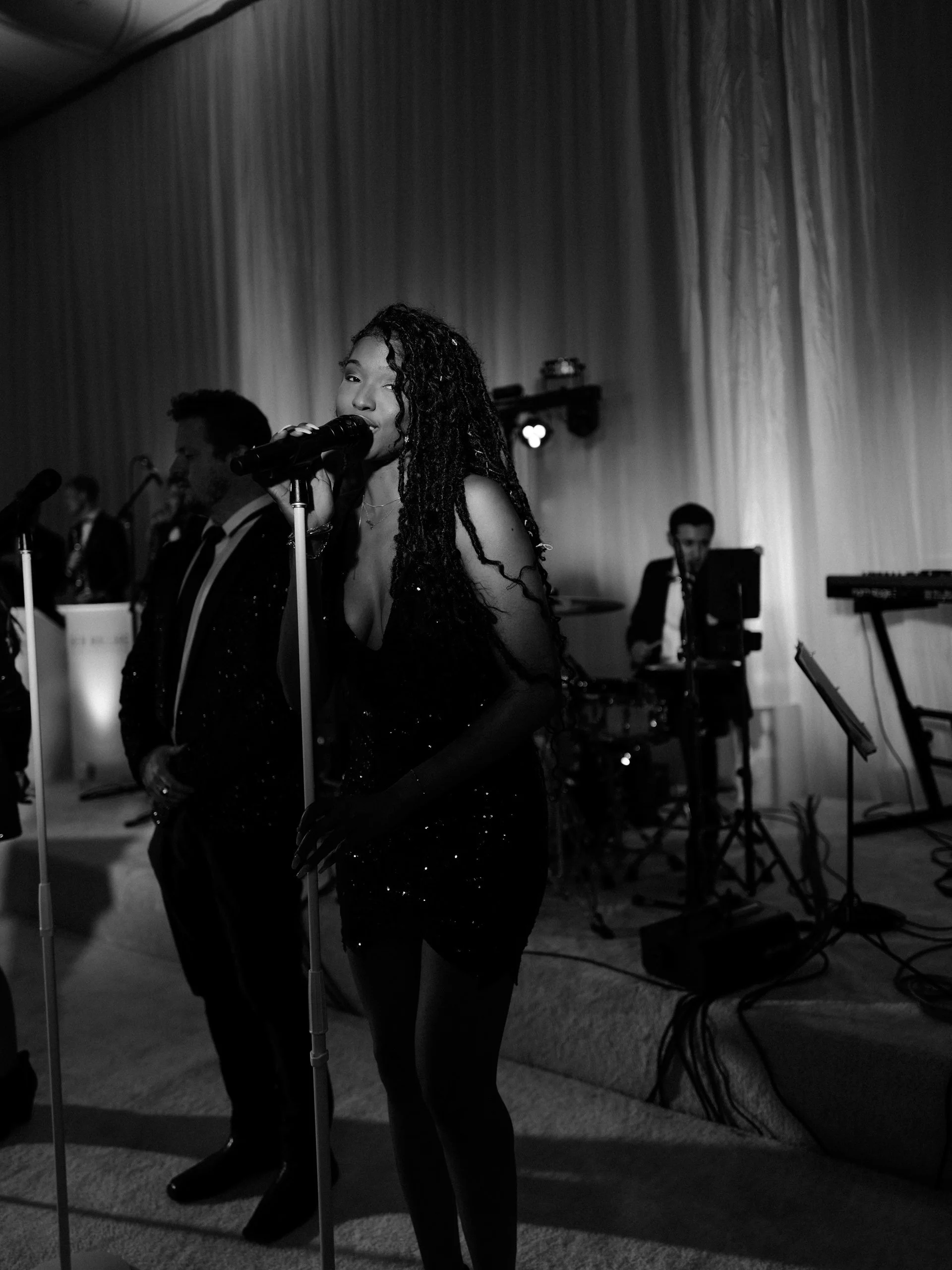 A female singer with long curly hair wearing a sparkly black dress, singing into a microphone on stage with a band in the background.