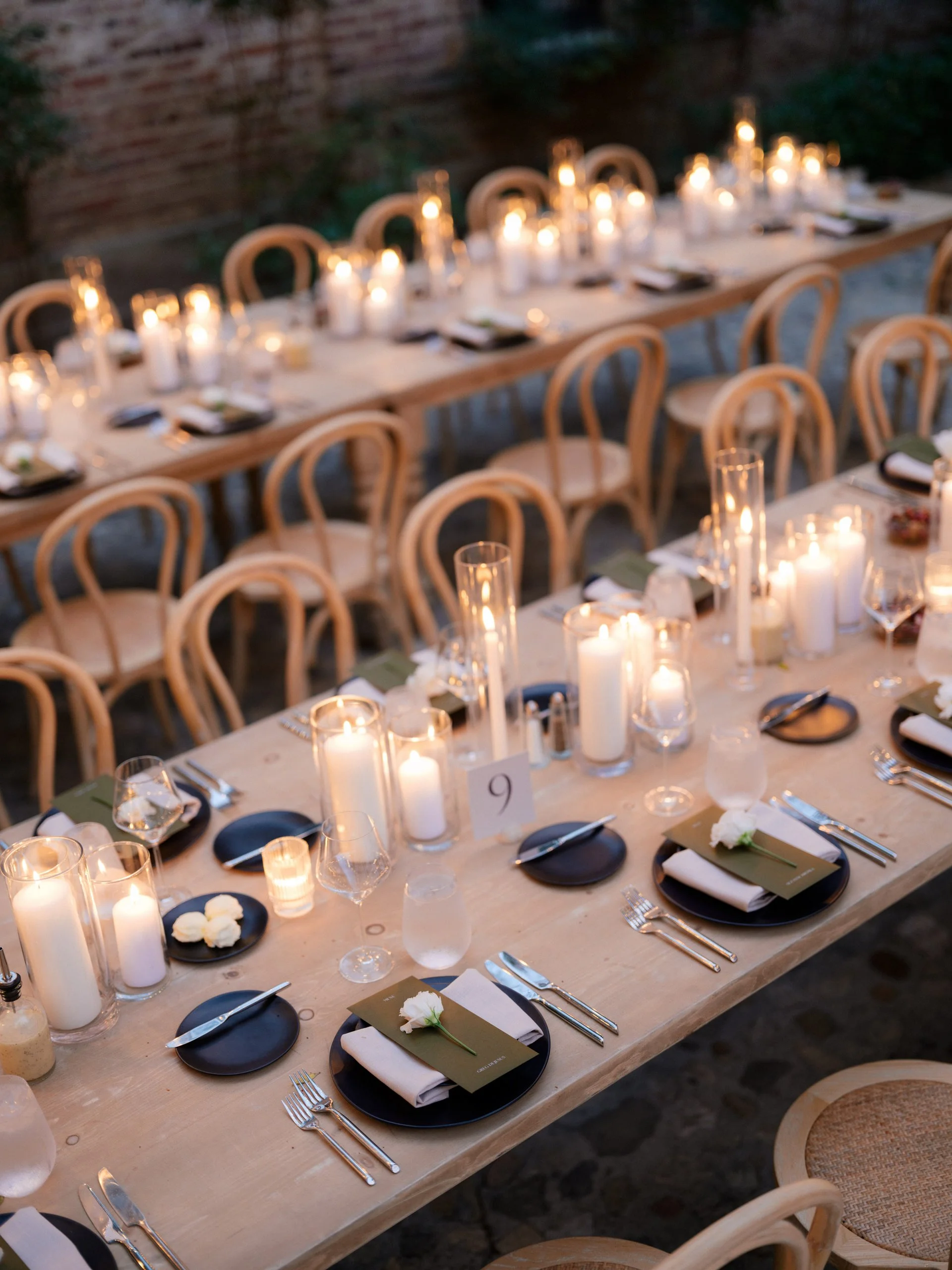 A long outdoor dinner table decorated with candles, wine glasses, black plates, cutlery, and napkins with white flowers, set for a formal event at dusk.