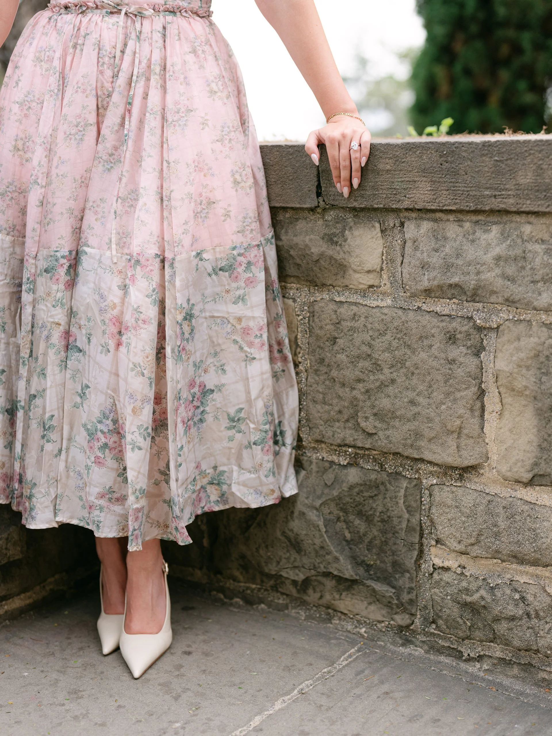 A woman wearing a pink floral skirt and white pointed high heels, resting her hand on a stone wall with a diamond ring and wearing a bracelet.
