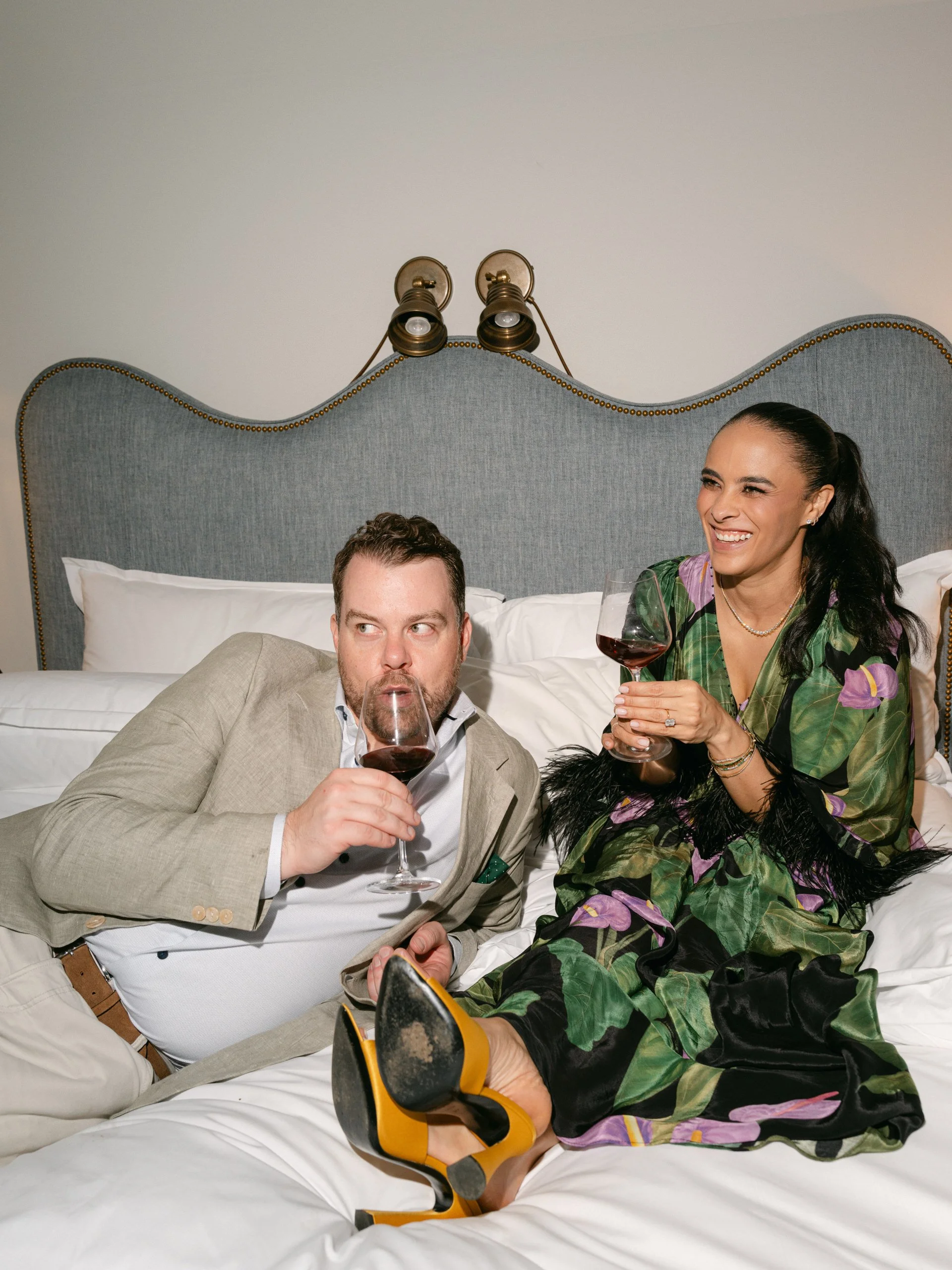 A man and woman sitting on a bed, drinking red wine and smiling. The woman is wearing a colorful dress with a floral pattern and yellow shoes, while the man is dressed in a beige suit.