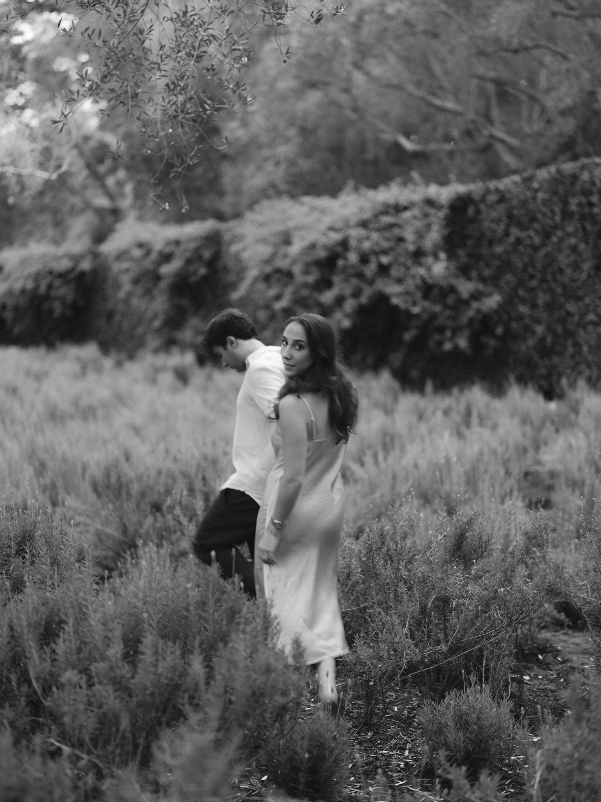 A black and white photo of a woman in a dress and a man in a shirt in a field of tall grass, with trees and bushes in the background.