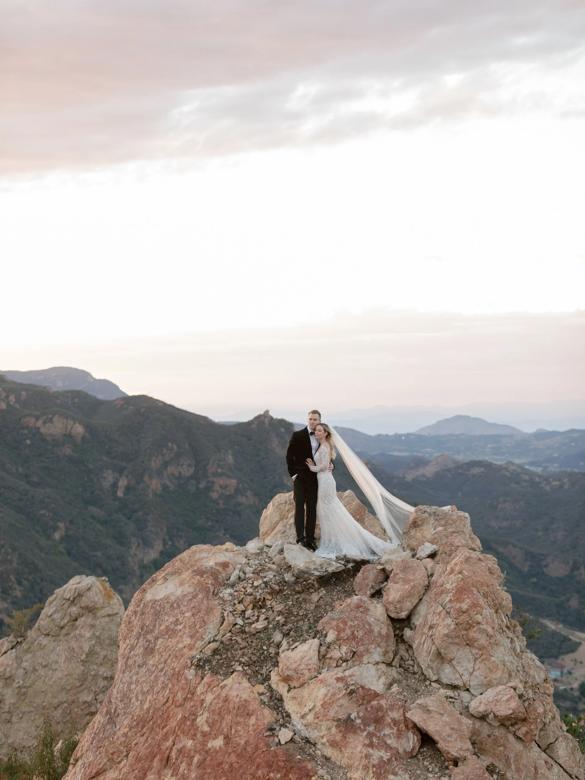 A bride and groom in wedding attire standing on a large rock formation in a mountainous landscape during sunset.