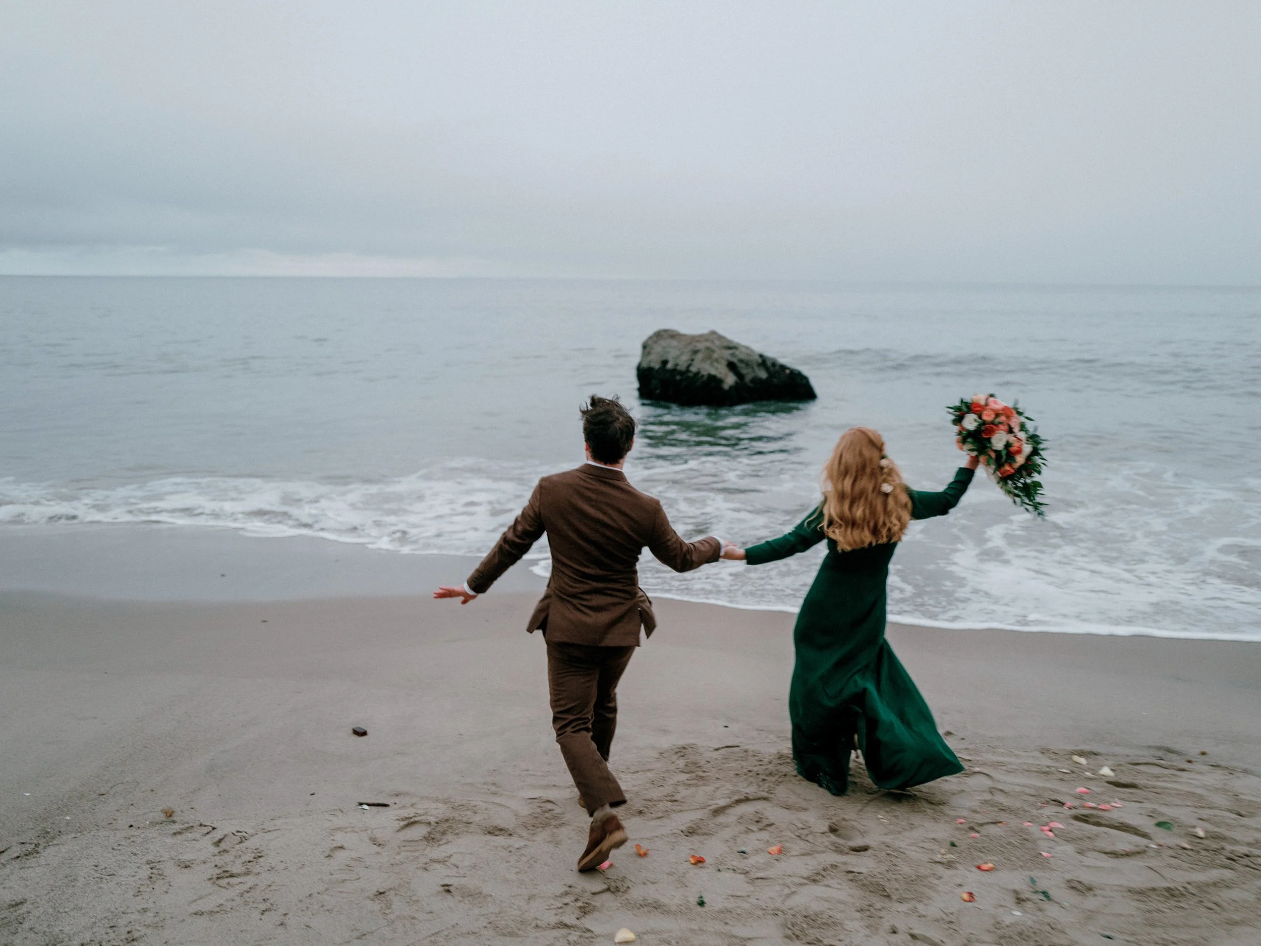 A couple in wedding attire holding hands and running on a beach with grey sand, with a large rock in the ocean and a cloudy sky in the background. The woman is holding a bouquet of flowers.