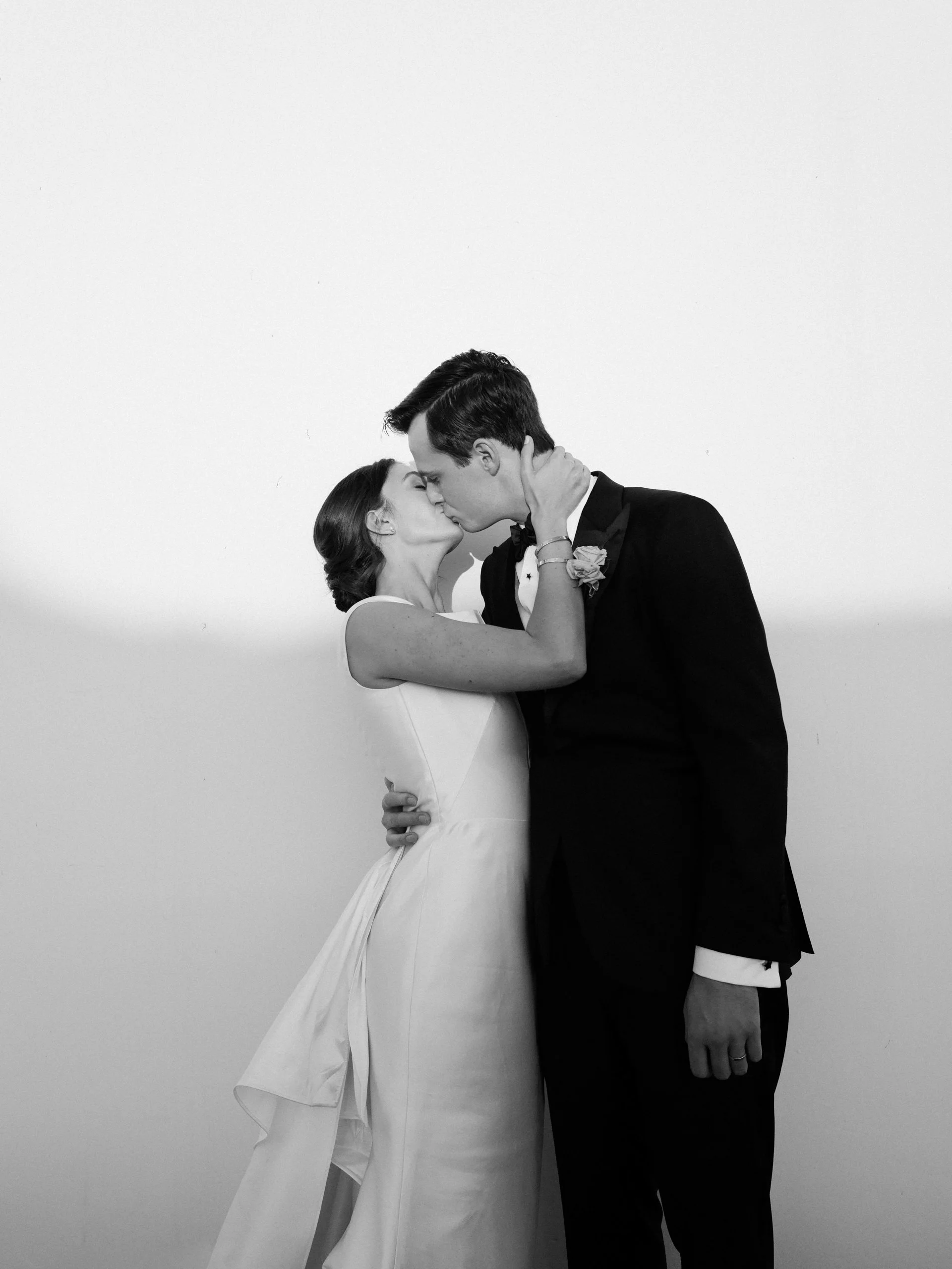 A black and white photo of a bride and groom sharing a kiss, with the bride holding the groom's face and the groom holding the bride's waist.