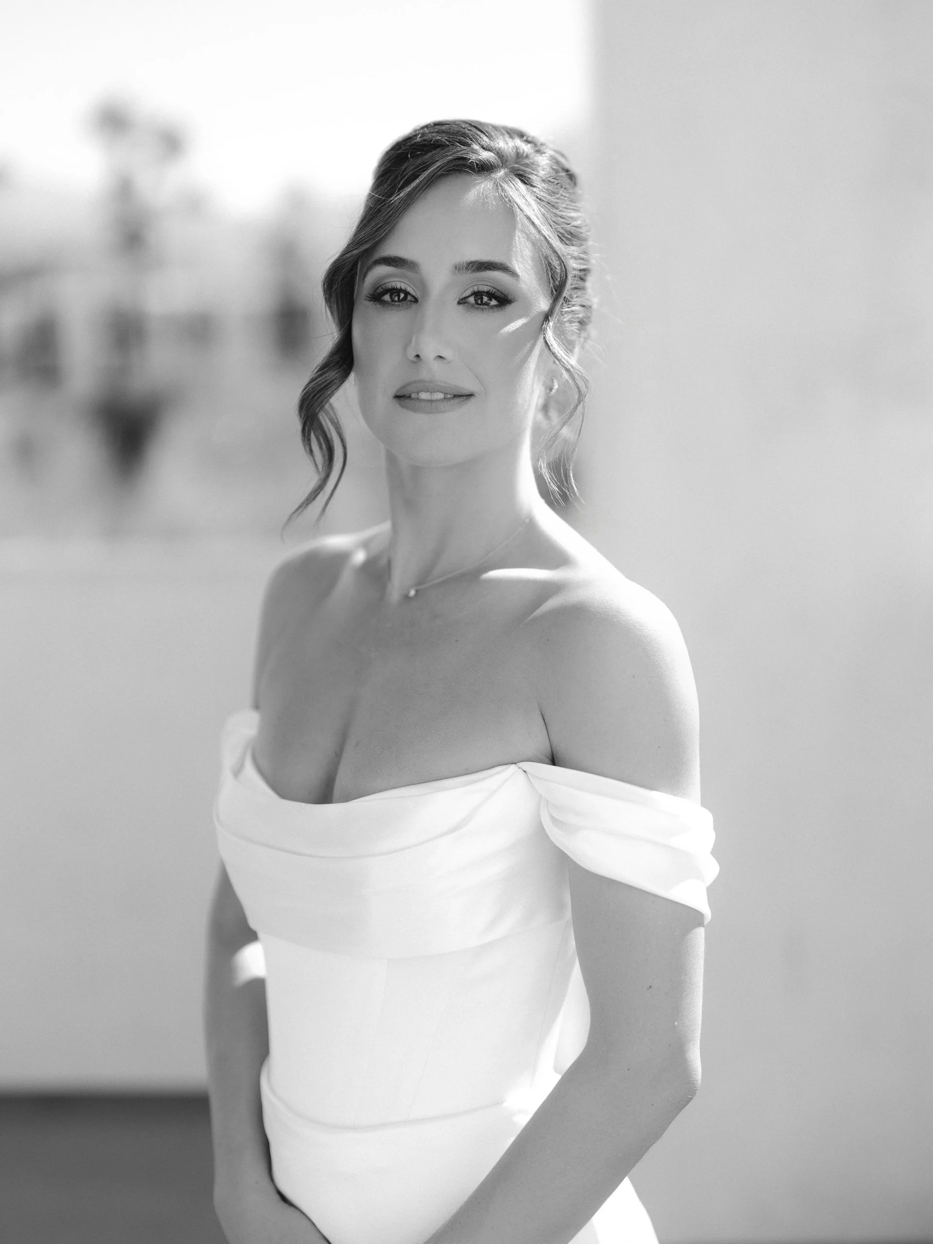 A black and white photo of a woman in an off-the-shoulder dress, with styled hair and makeup, standing outdoors on a sunny day.
