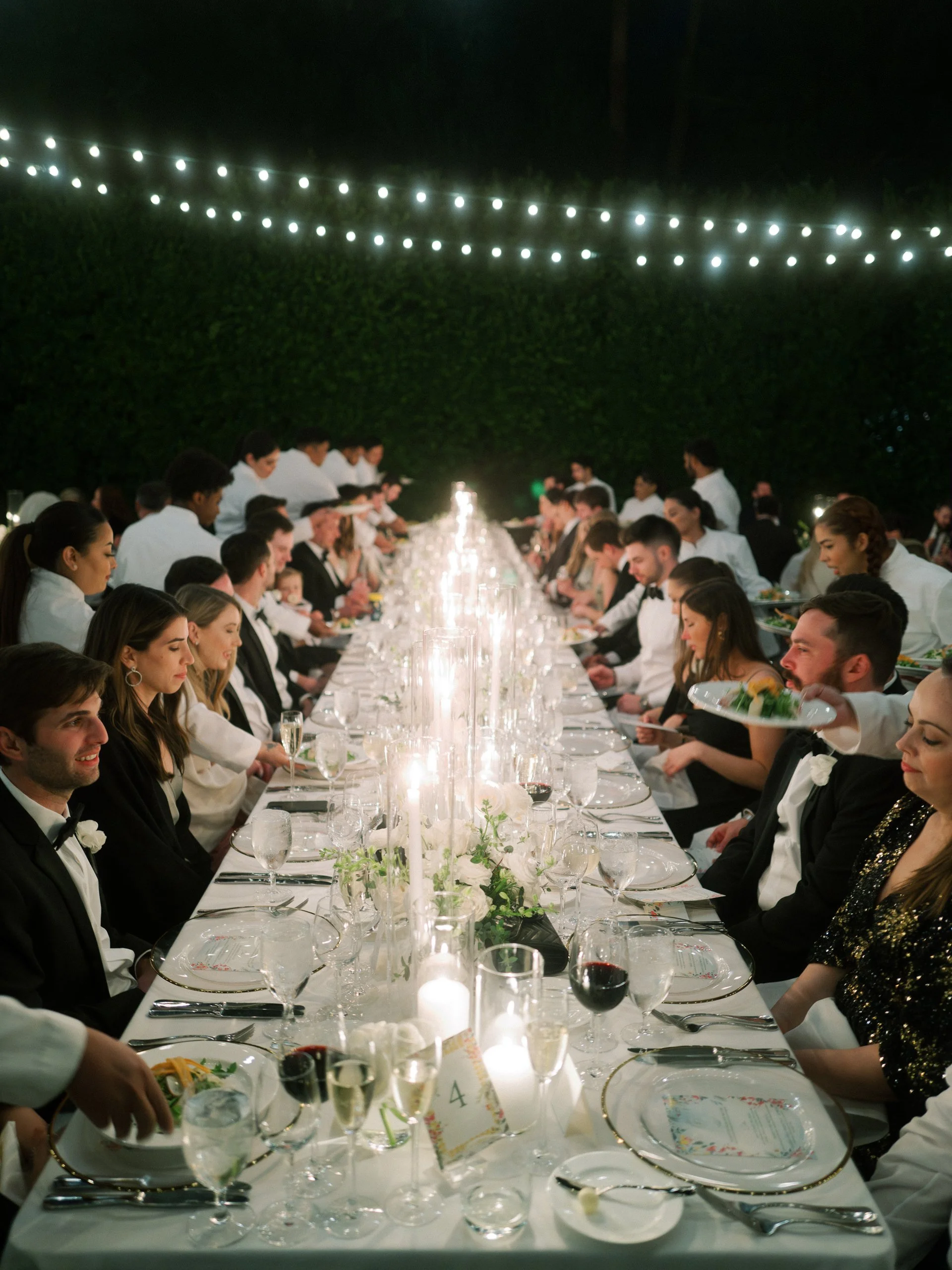 A wedding reception with guests seated at a long, elegantly set table outdoors at night, decorated with white flowers and candles, with string lights above.