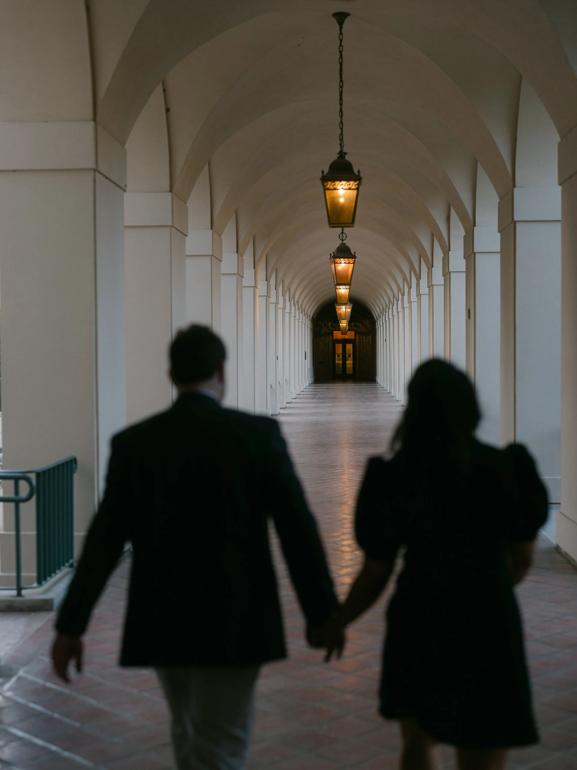 A man and woman in formal attire walking hand in hand through an arched walkway with hanging lanterns.