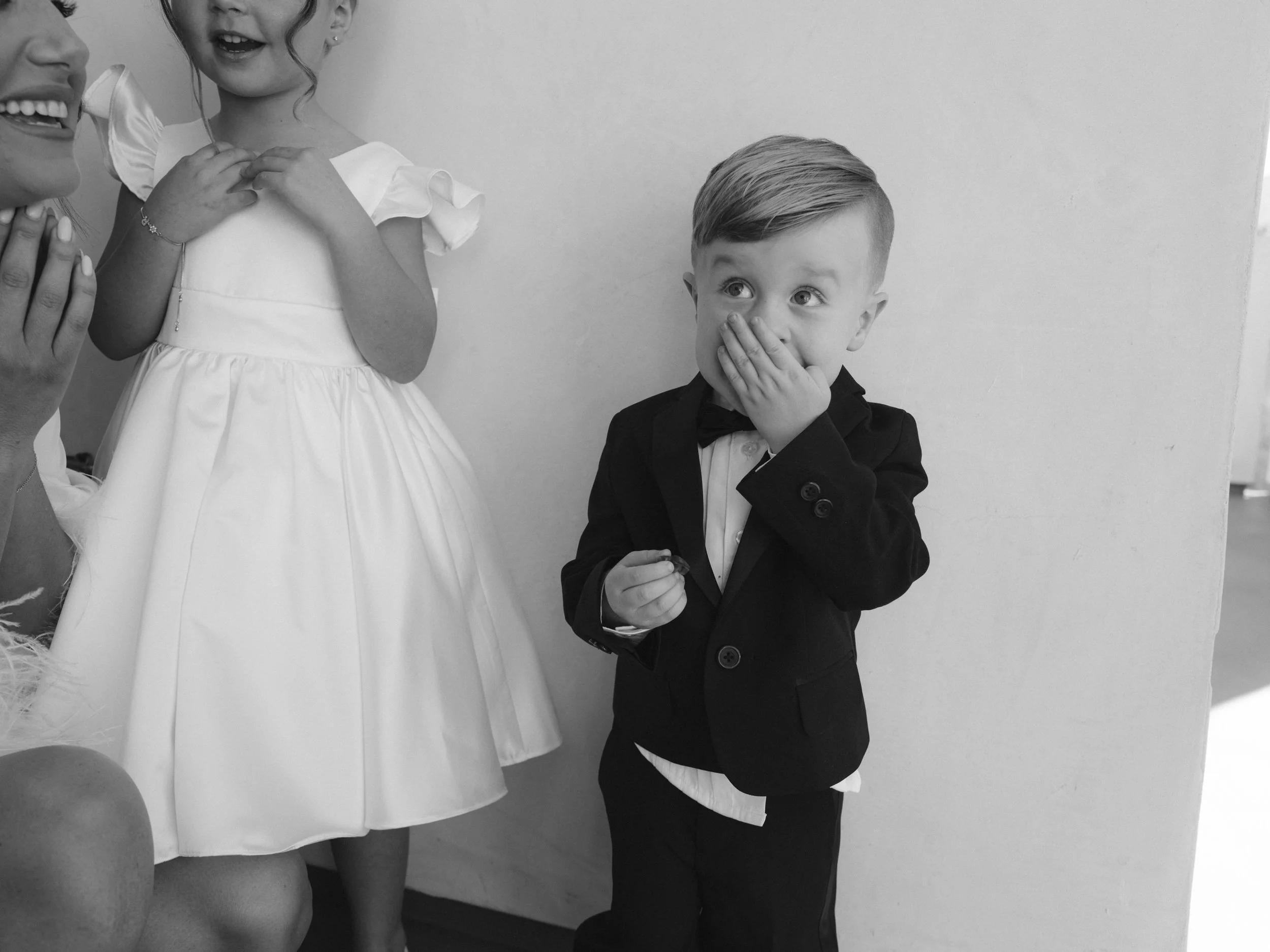 A young boy in a suit with a bow tie covering his mouth in surprise, standing next to a girl in a white dress and an adult woman smiling.
