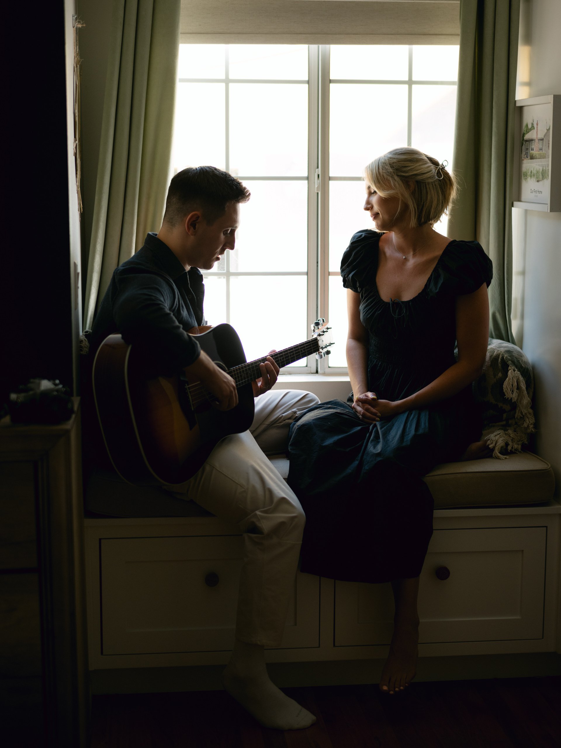 A man playing guitar and a woman sitting on a window seat, facing each other, in a cozy room with natural light and green curtains.