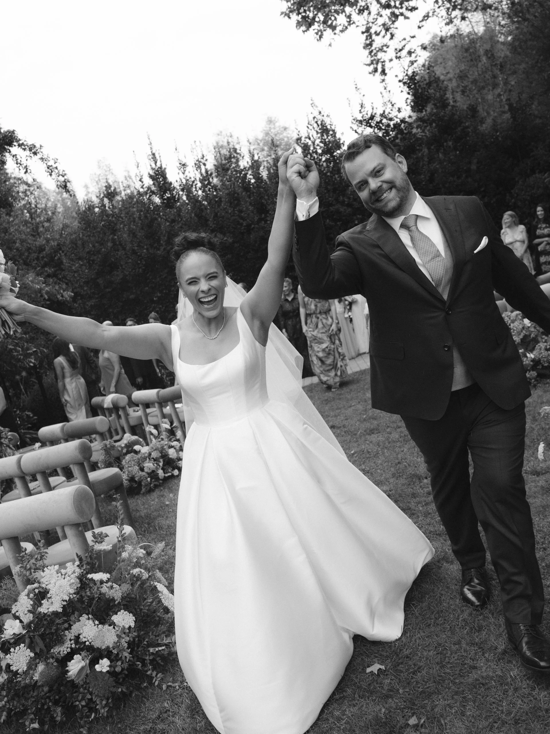 Bride and groom celebrating at their wedding outdoors, holding hands and smiling.