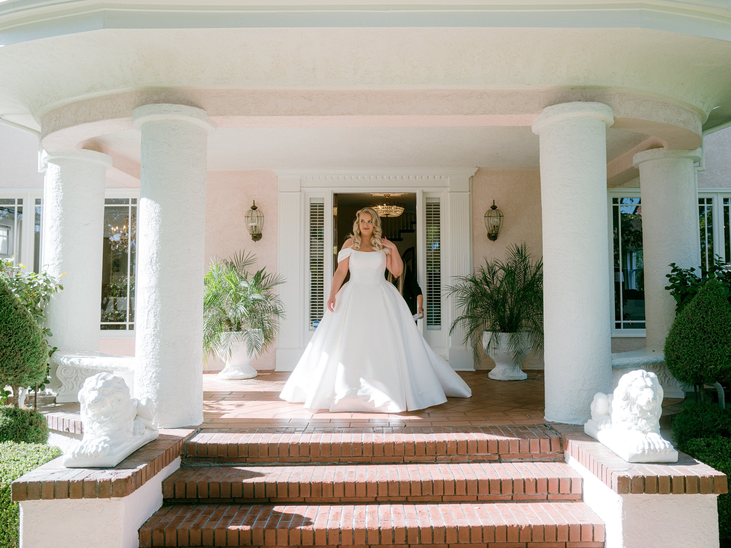 A bride in a white wedding gown standing on the steps of a house with white columns and potted plants.