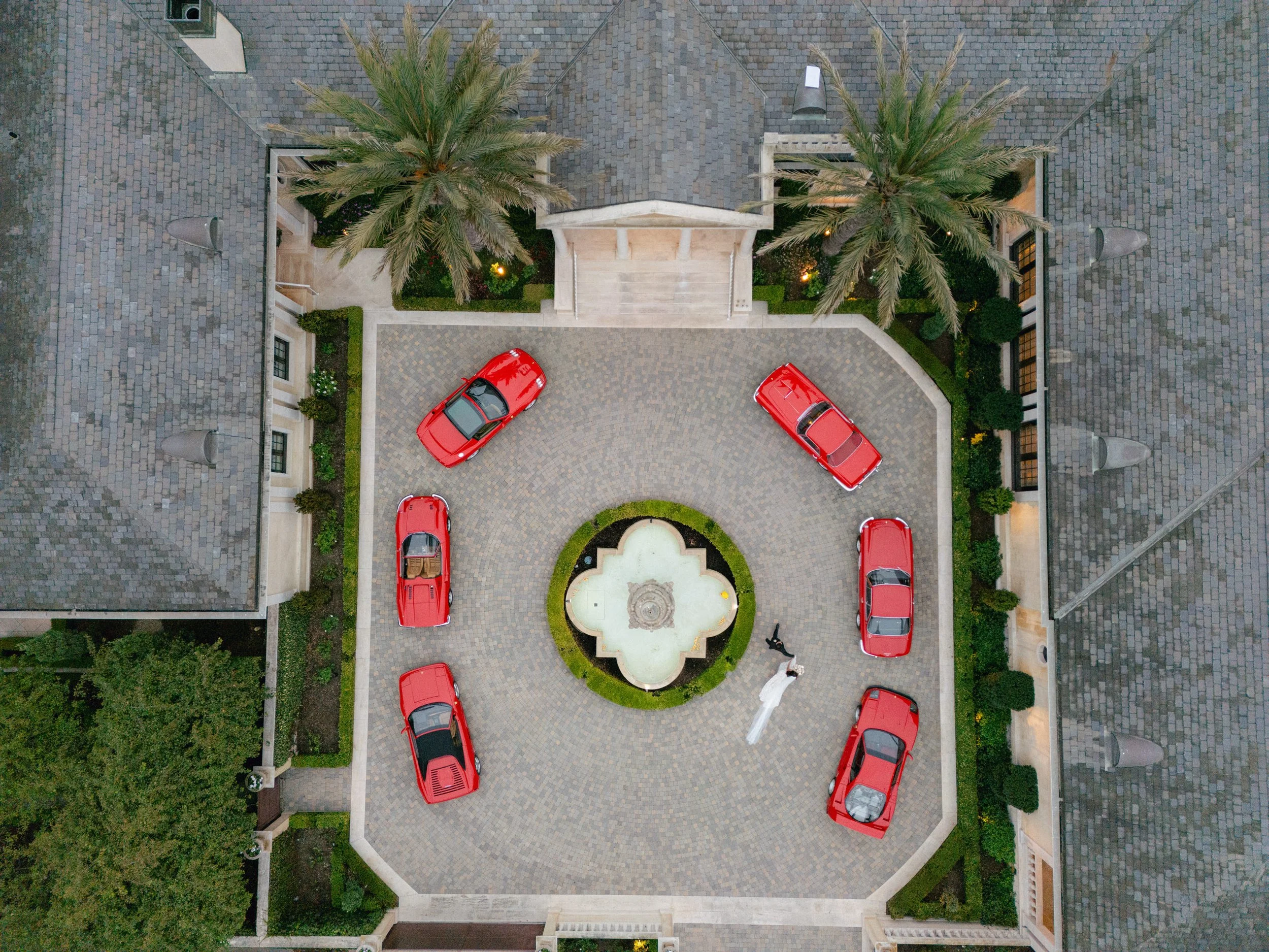 An overhead view of a courtyard with seven red cars parked in a circular pattern around a fountain, with palm trees and buildings surrounding the area.