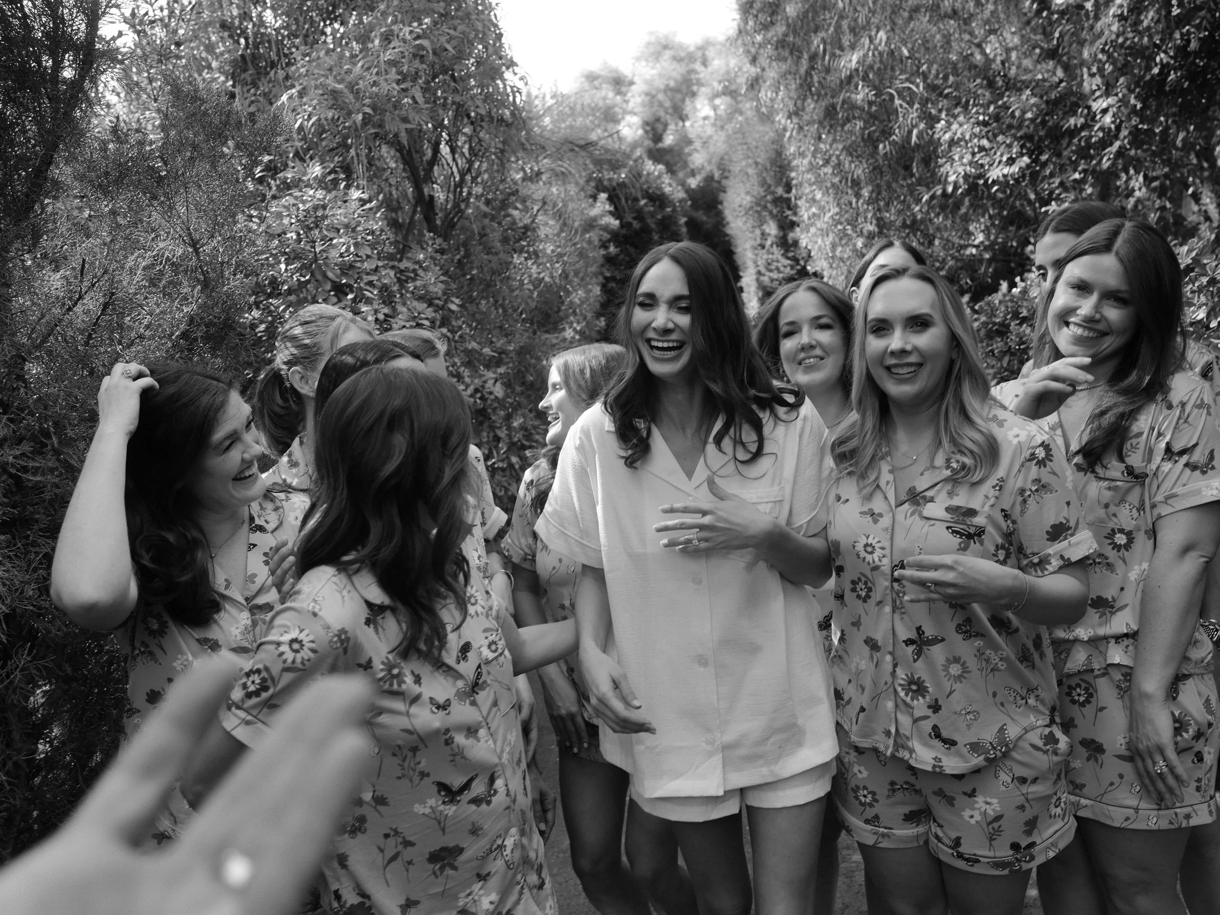 Group of women in pajamas laughing and talking outdoors among trees and bushes.