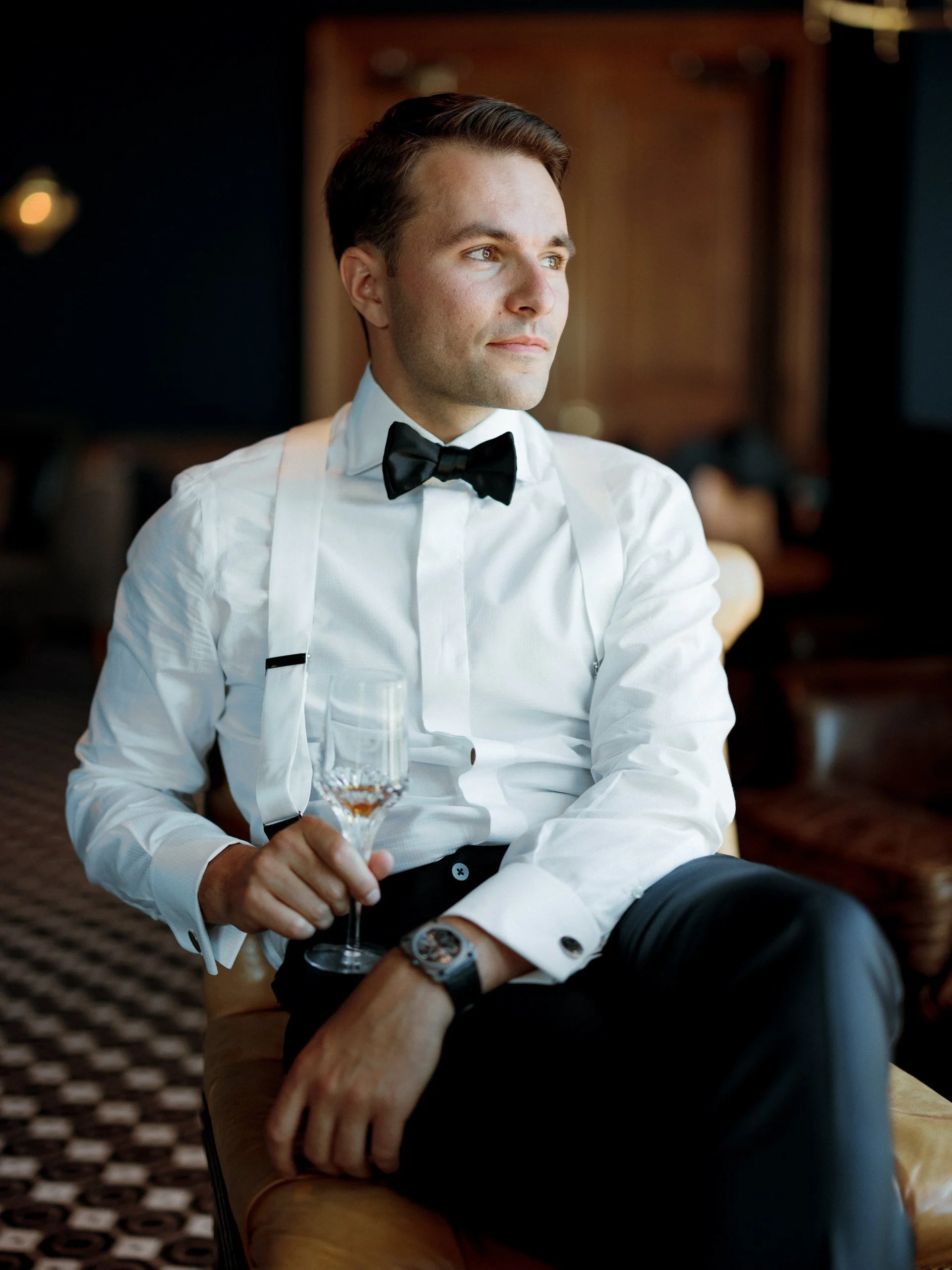 A young man in formal attire, wearing a white dress shirt, black bow tie, and suspenders, sitting on a yellow couch in a luxurious setting holding a glass of champagne.