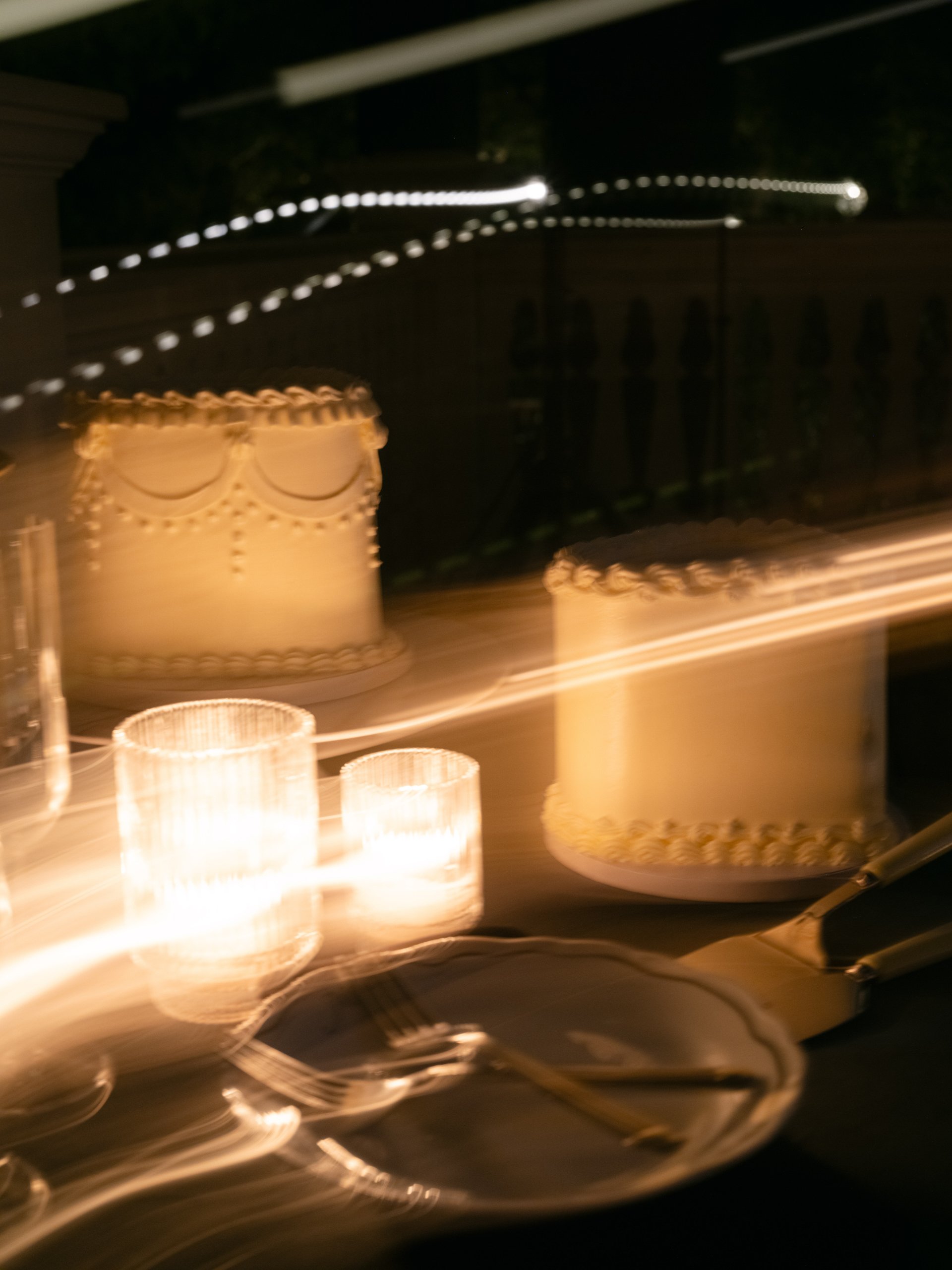 Two decorated cakes on a table at night, with candles and utensils.