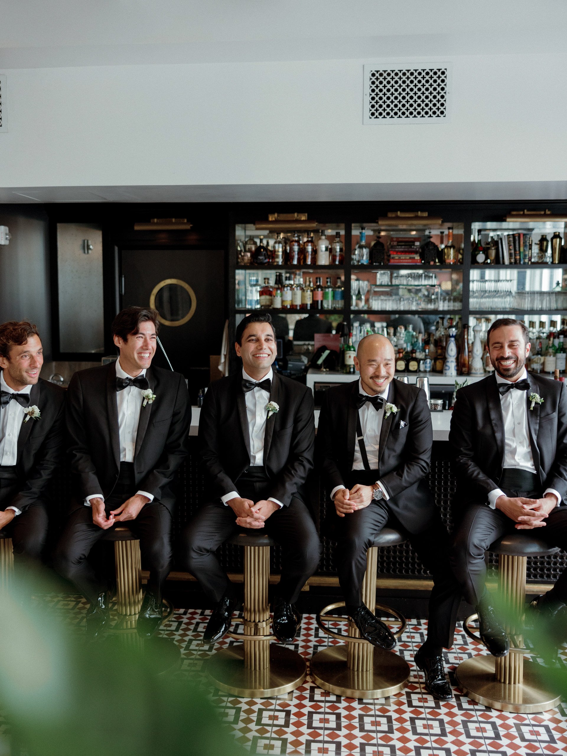 Five men dressed in black tuxedos with bow ties, sitting on bar stools in a bar or restaurant, smiling and laughing together.