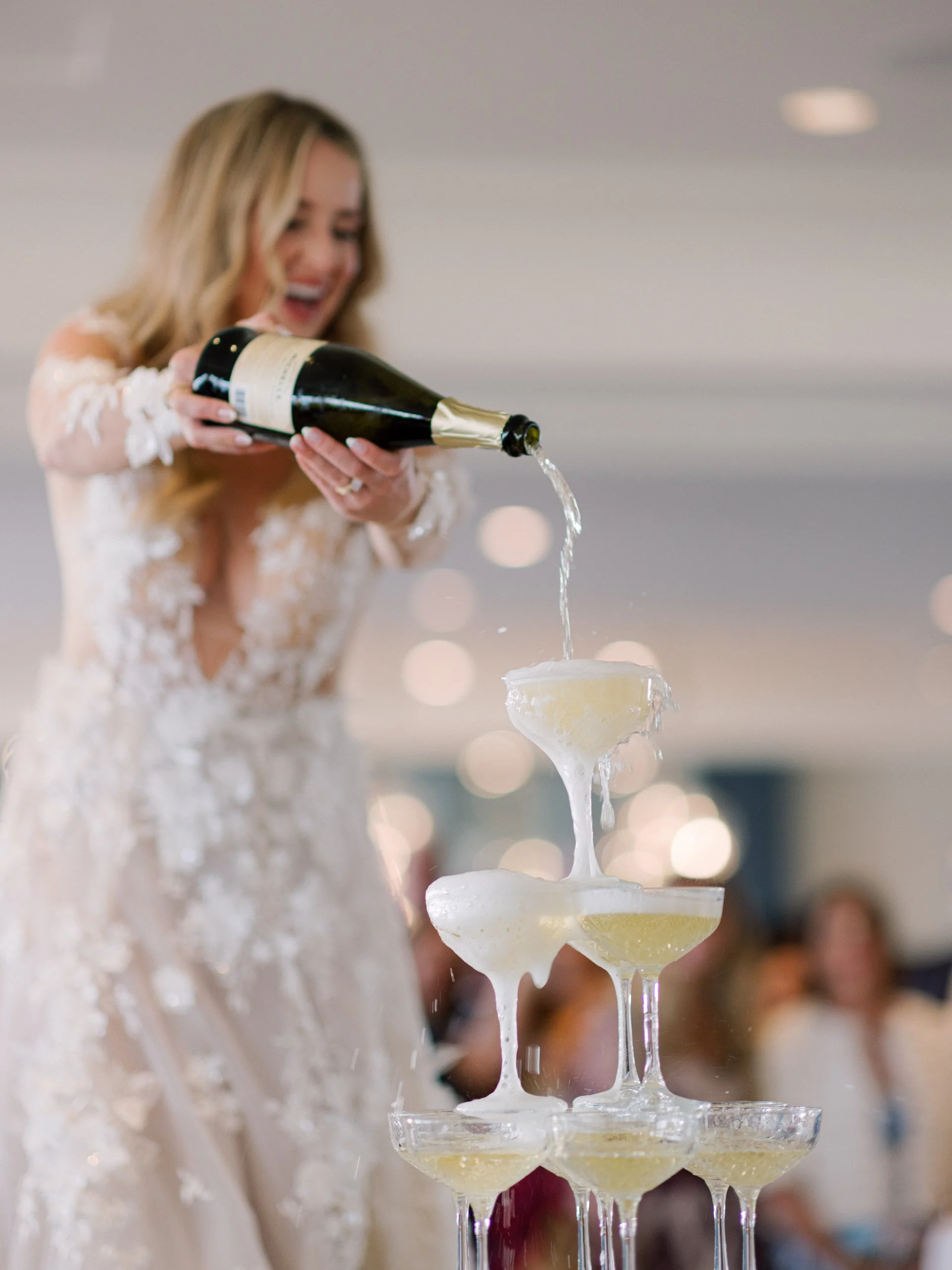 A woman in a wedding dress is pouring champagne into a pyramid of glasses at a celebration.