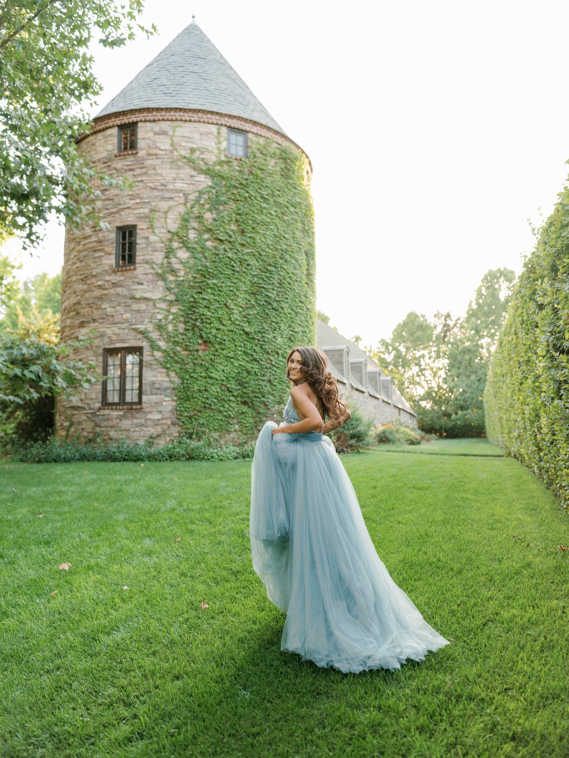 A woman in a flowing blue dress walking in a lush green lawn near an old stone tower covered with ivy, with a bright sky in the background.