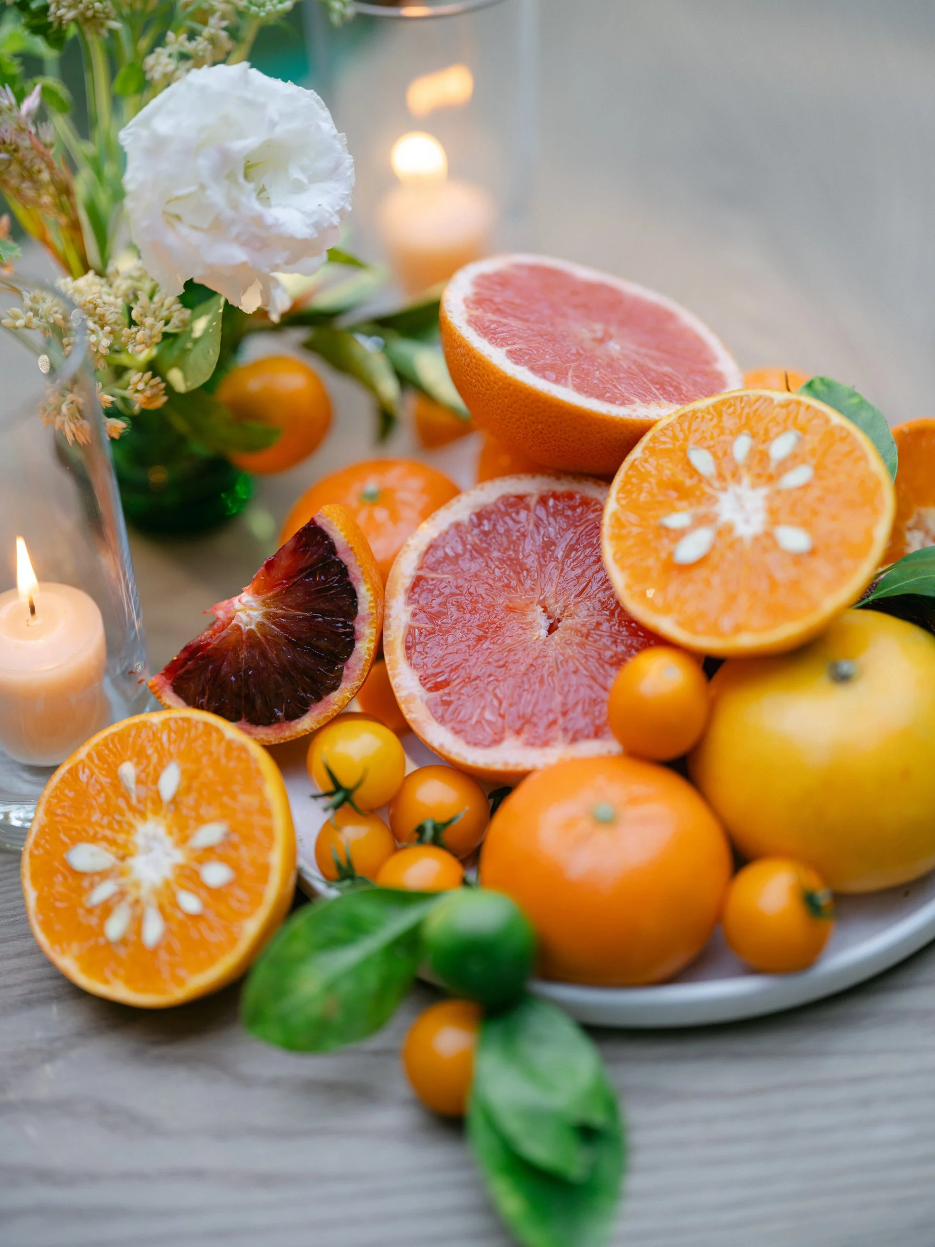 A plate with halved grapefruits, oranges, and yellow cherry tomatoes, surrounded by green leaves, a white flower, and lit candles in the background.