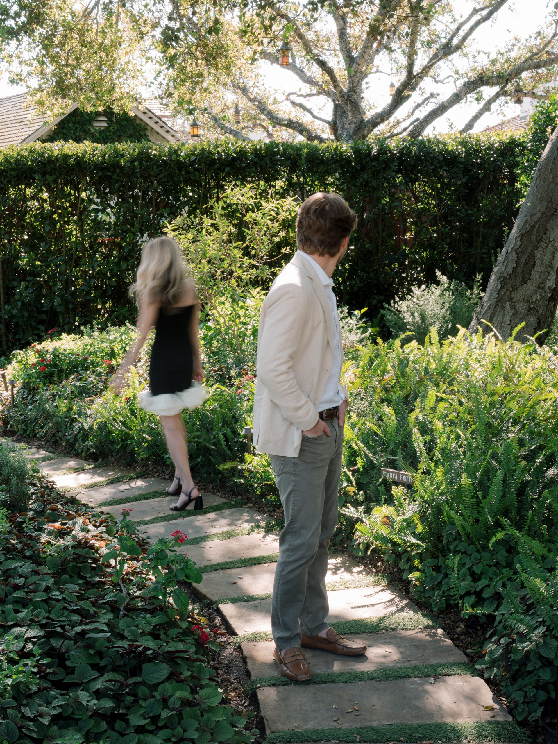 A man in a cream blazer and gray pants stands on a stone pathway in a lush garden, looking at the plants. A woman in a black dress and high heels walks away along the pathway, surrounded by greenery and trees.