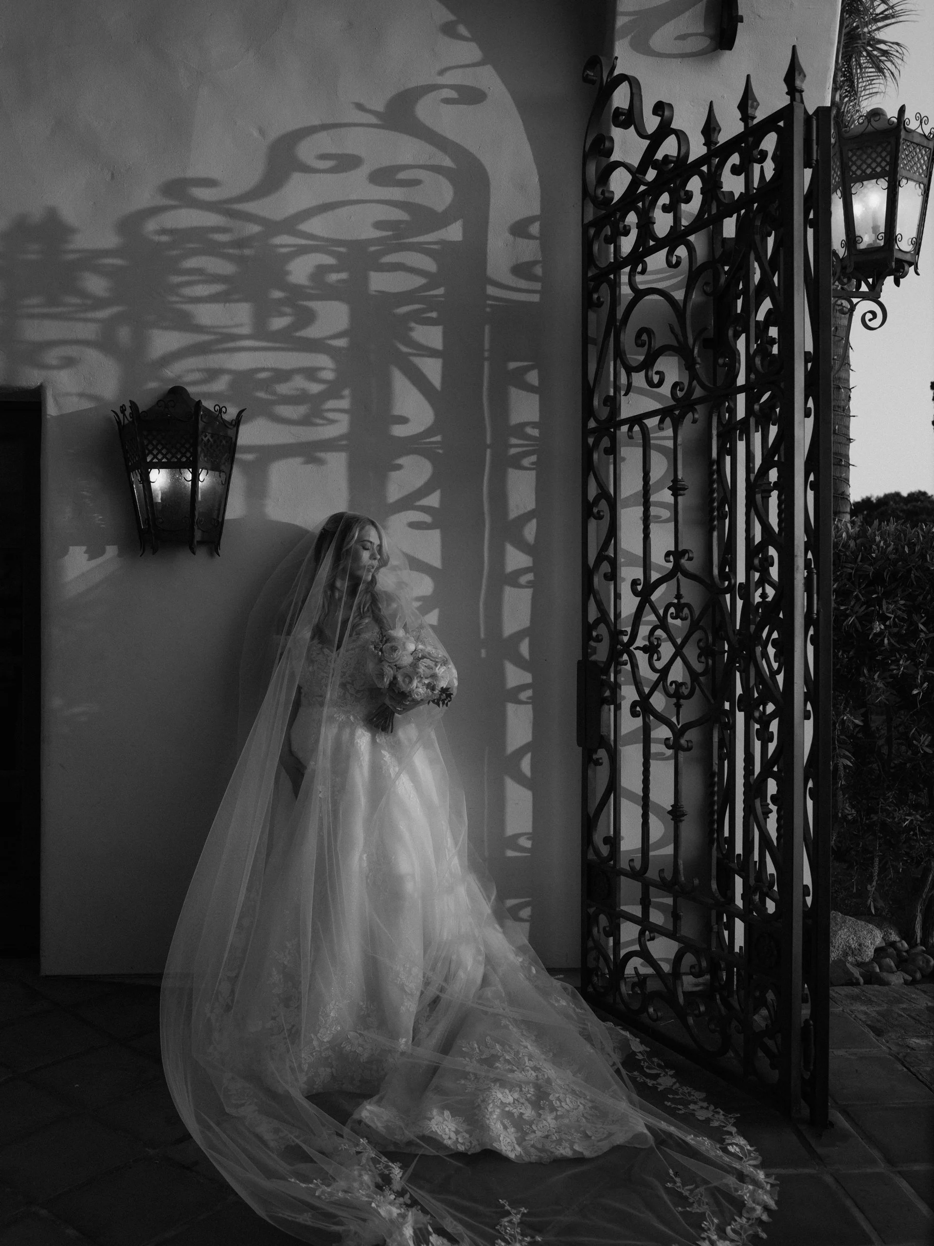A bride in a wedding gown with a veil holding a bouquet, standing near an ornate wrought-iron gate, with shadow patterns cast on the wall behind her.