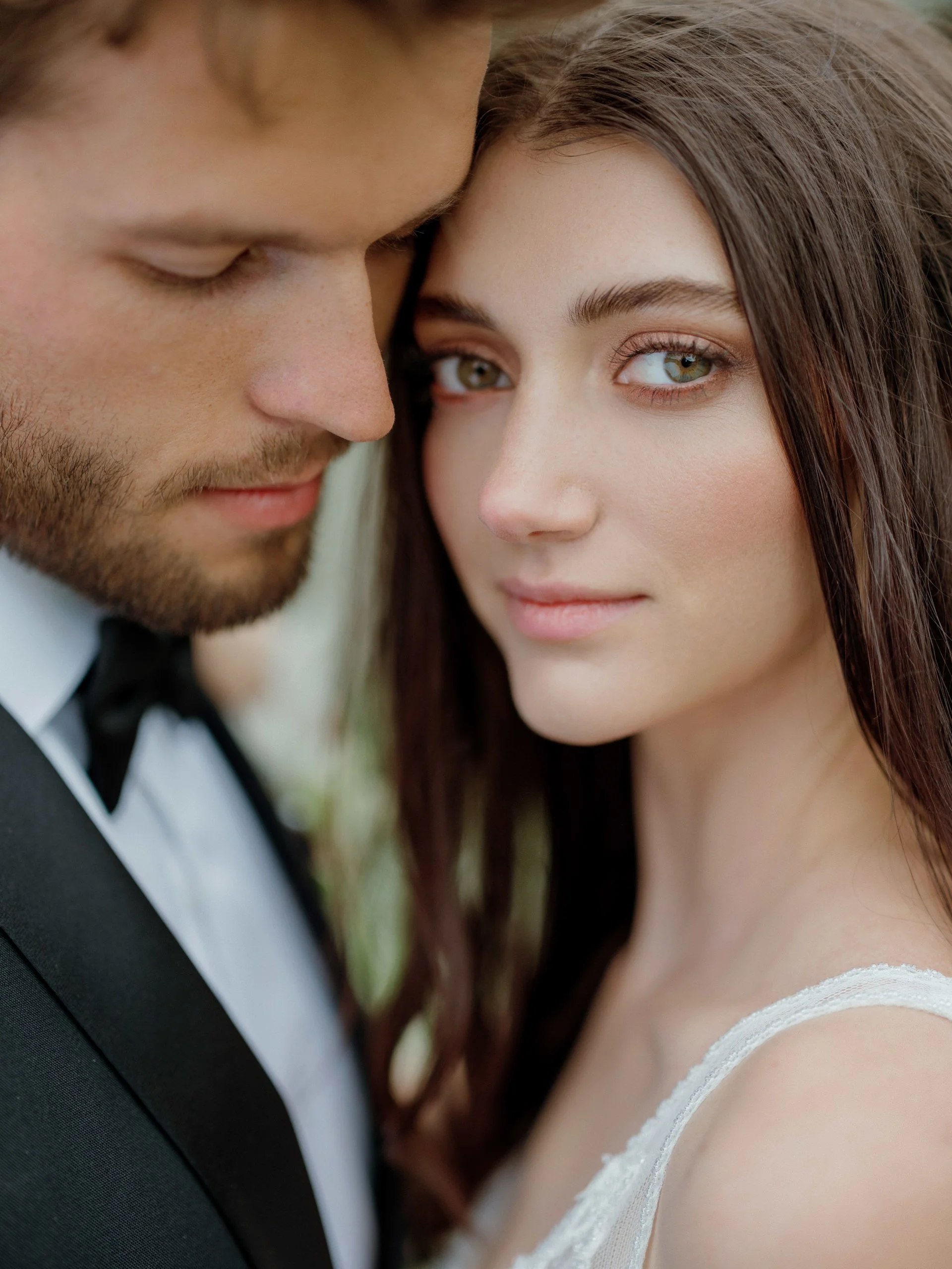 Close-up of a groom and bride with their faces close together, both looking at the camera. The groom is wearing a tuxedo with a bow tie, and the bride has long brown hair and is wearing a white dress.