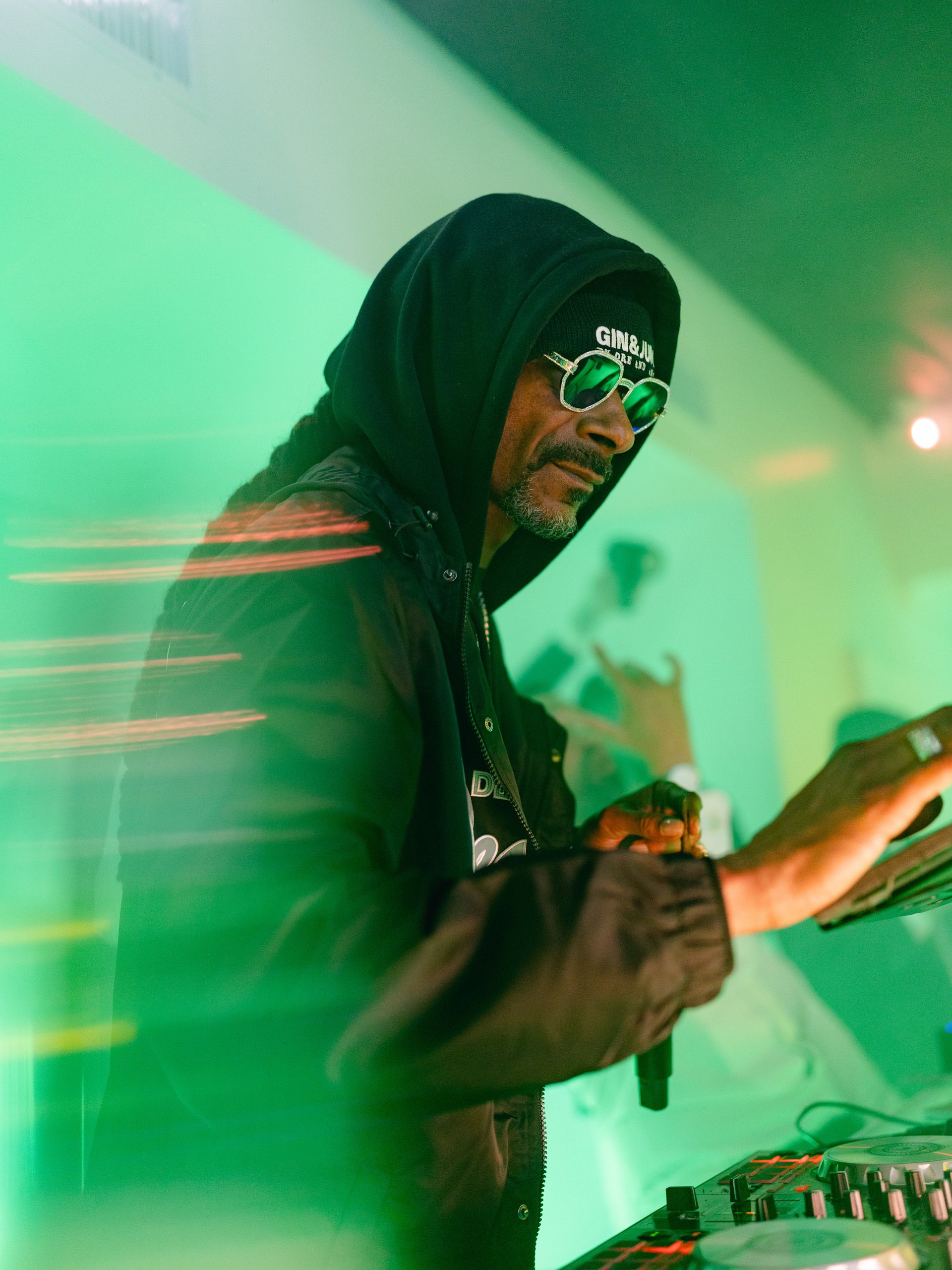 A DJ wearing sunglasses, a black hoodie, and a black jacket, standing behind DJ equipment in a green-lit environment.