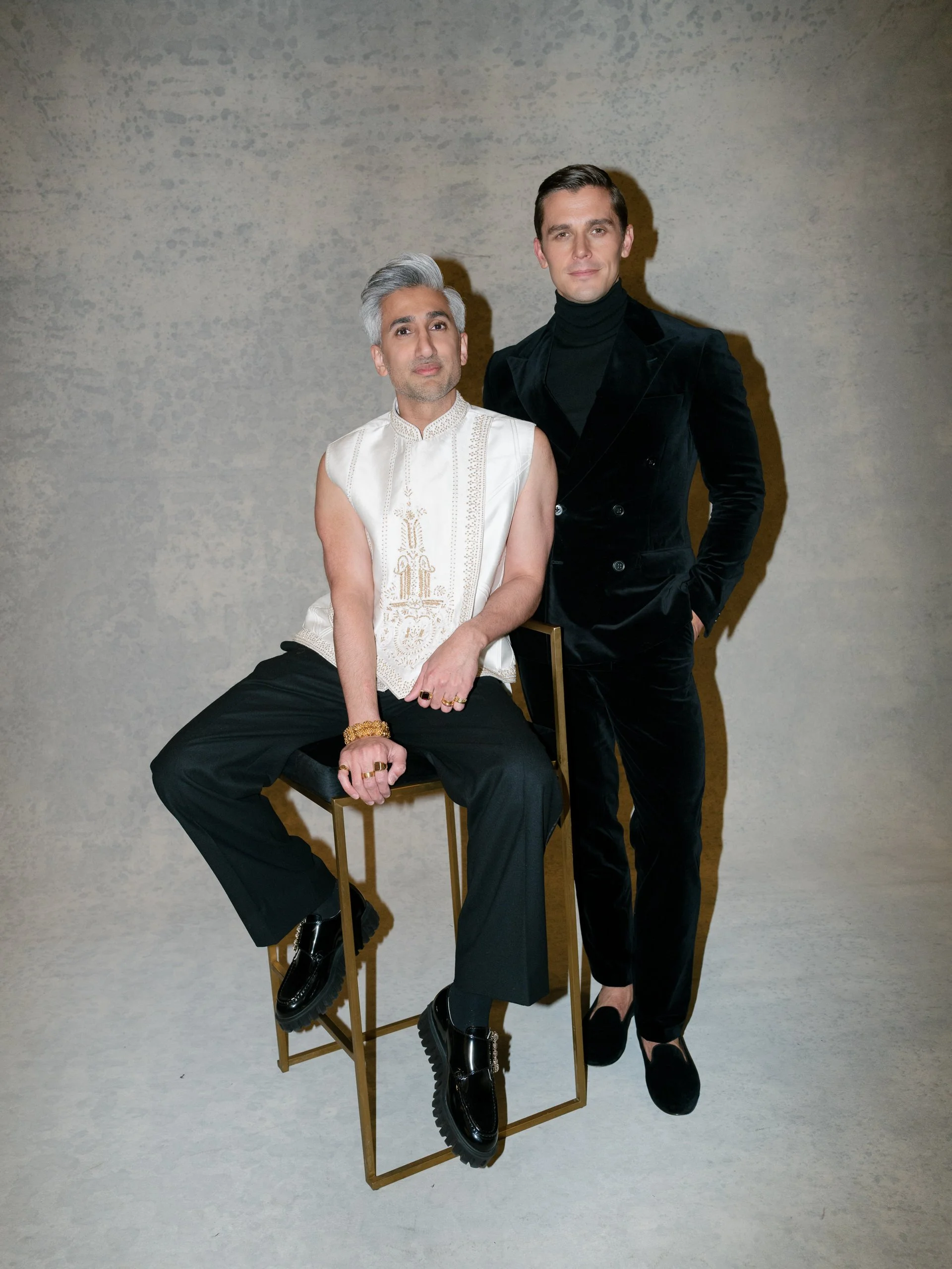 Two men, one seated on a chair and the other standing, posing against a plain textured background. The seated man has gray hair, wearing a sleeveless white embroidered top, black trousers, and black shiny shoes with chunky soles. He has jewelry on hi
