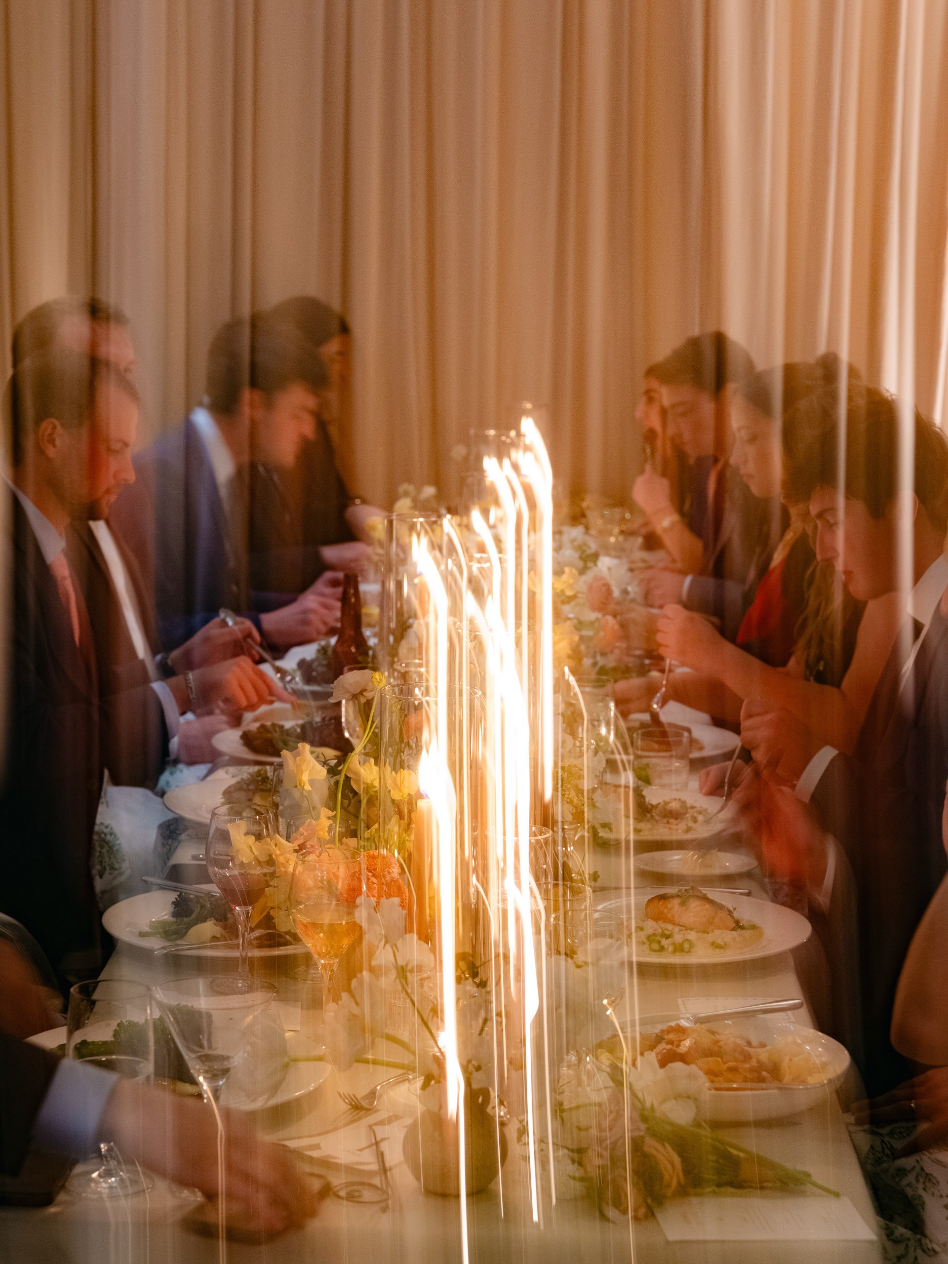 People seated at a long dining table, viewed through a glass with reflections or light streaks.