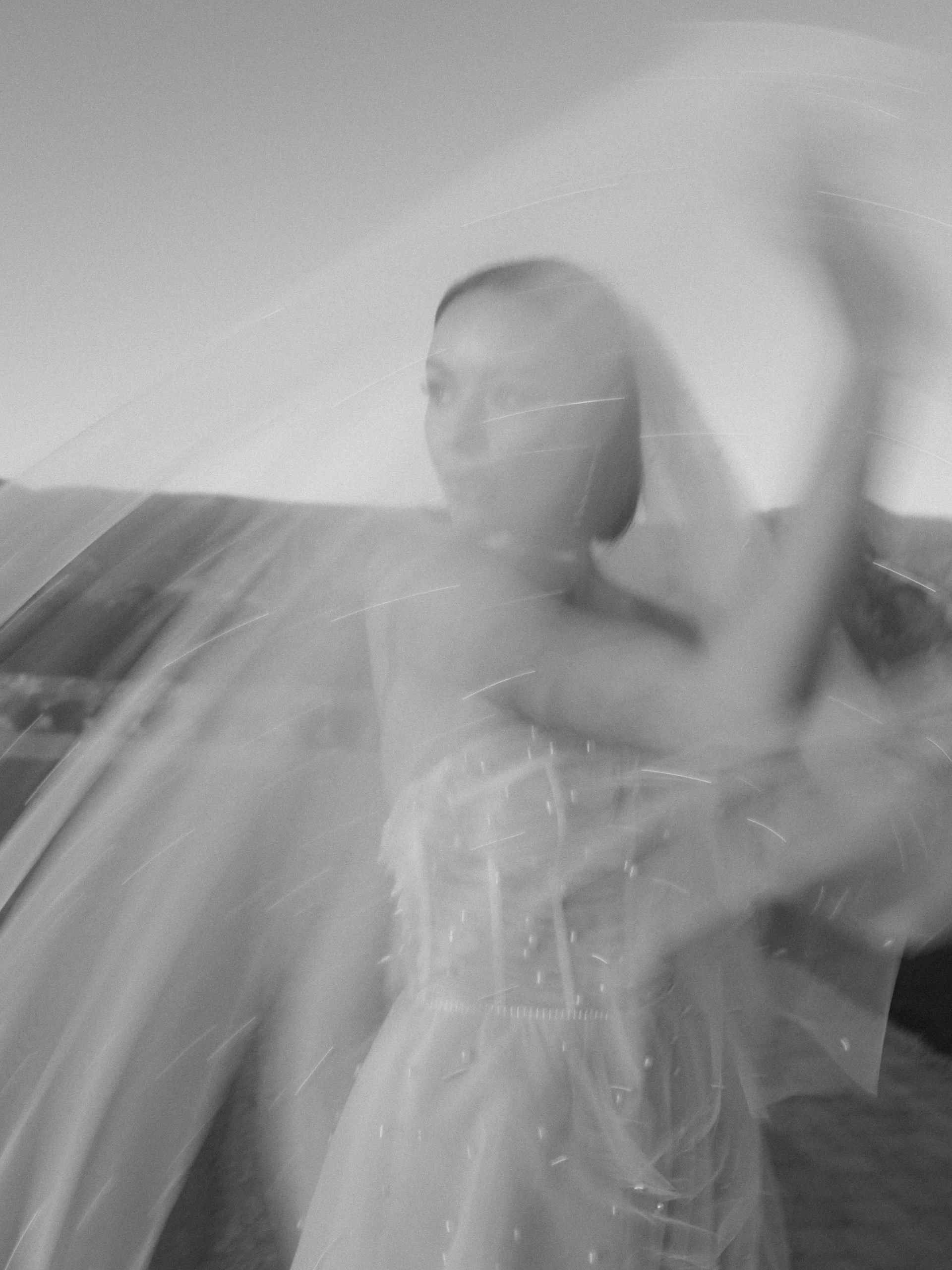 Blurred black and white photo of a woman taking a selfie through a glass surface, creating a reflection and motion blur effect.