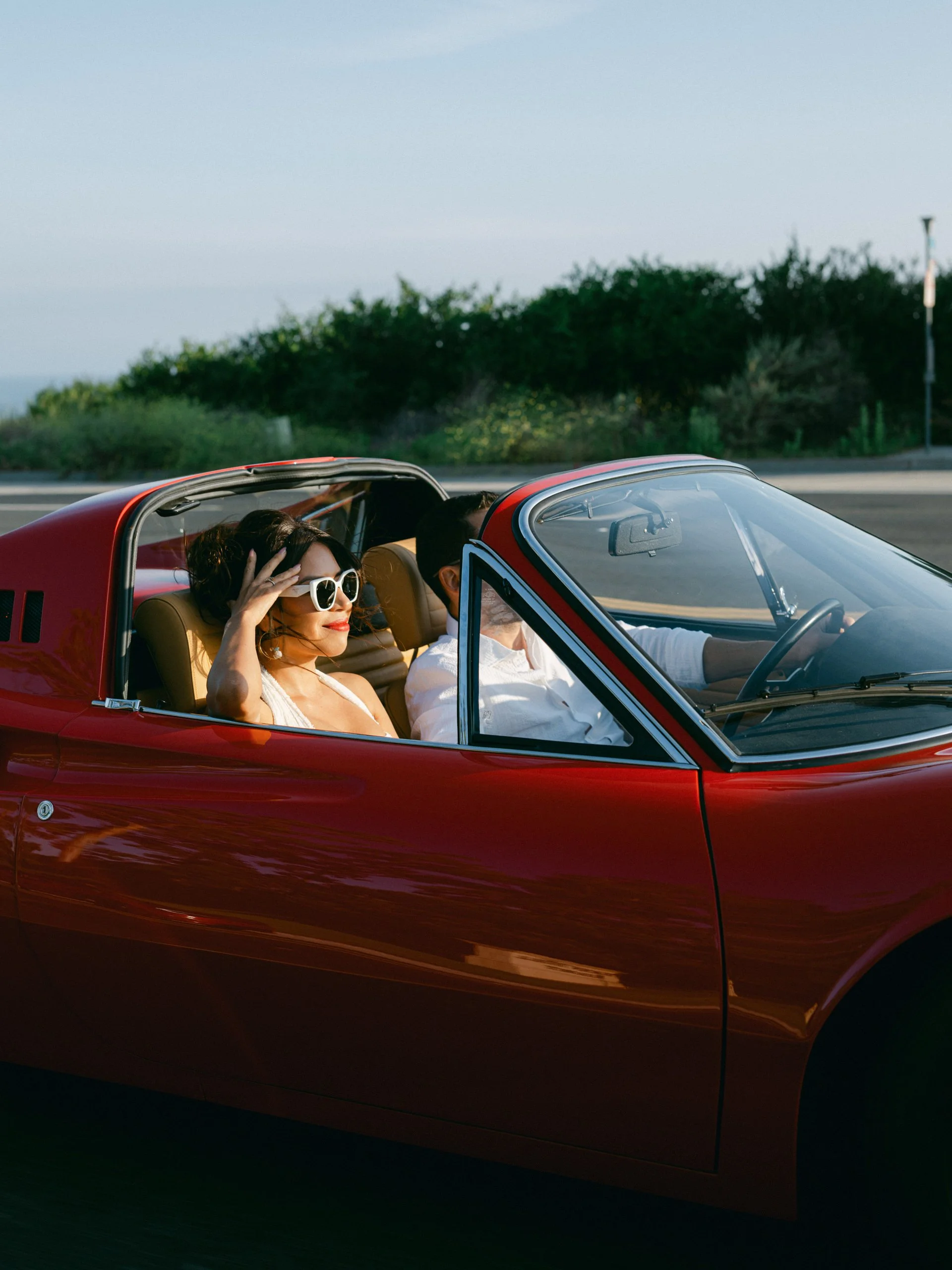 A woman wearing white sunglasses and a white top, sitting in a red convertible car, smiling and enjoying a sunny day with her hand on her head, while a man drives the car.
