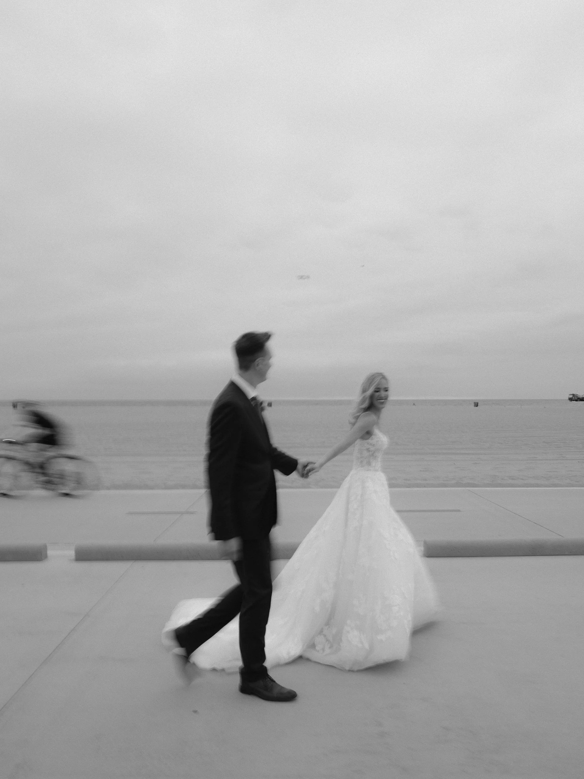 Black and white photo of a bride and groom holding hands by the seaside, with a cyclist blurred in motion in the background.