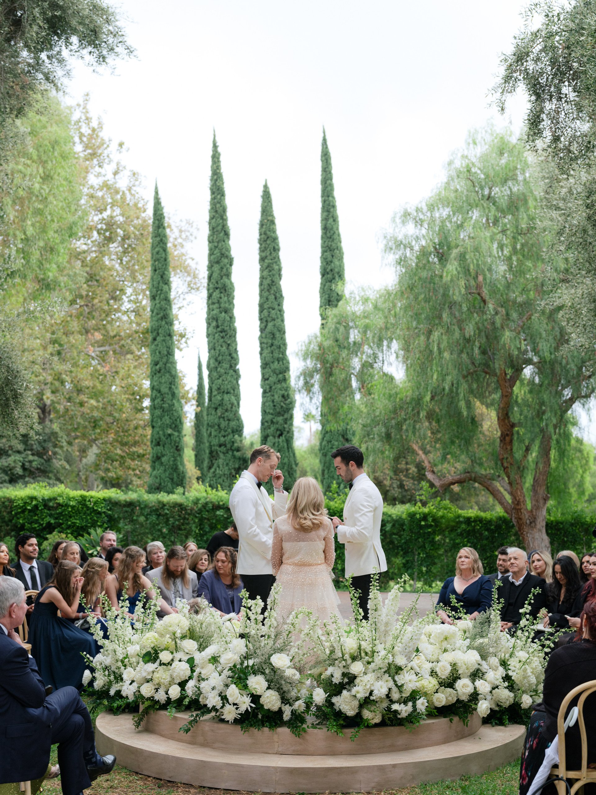 A wedding ceremony outdoors with a couple exchanging vows, officiant, and seated guests, surrounded by green trees and tall cypress trees, decorated with white flowers.