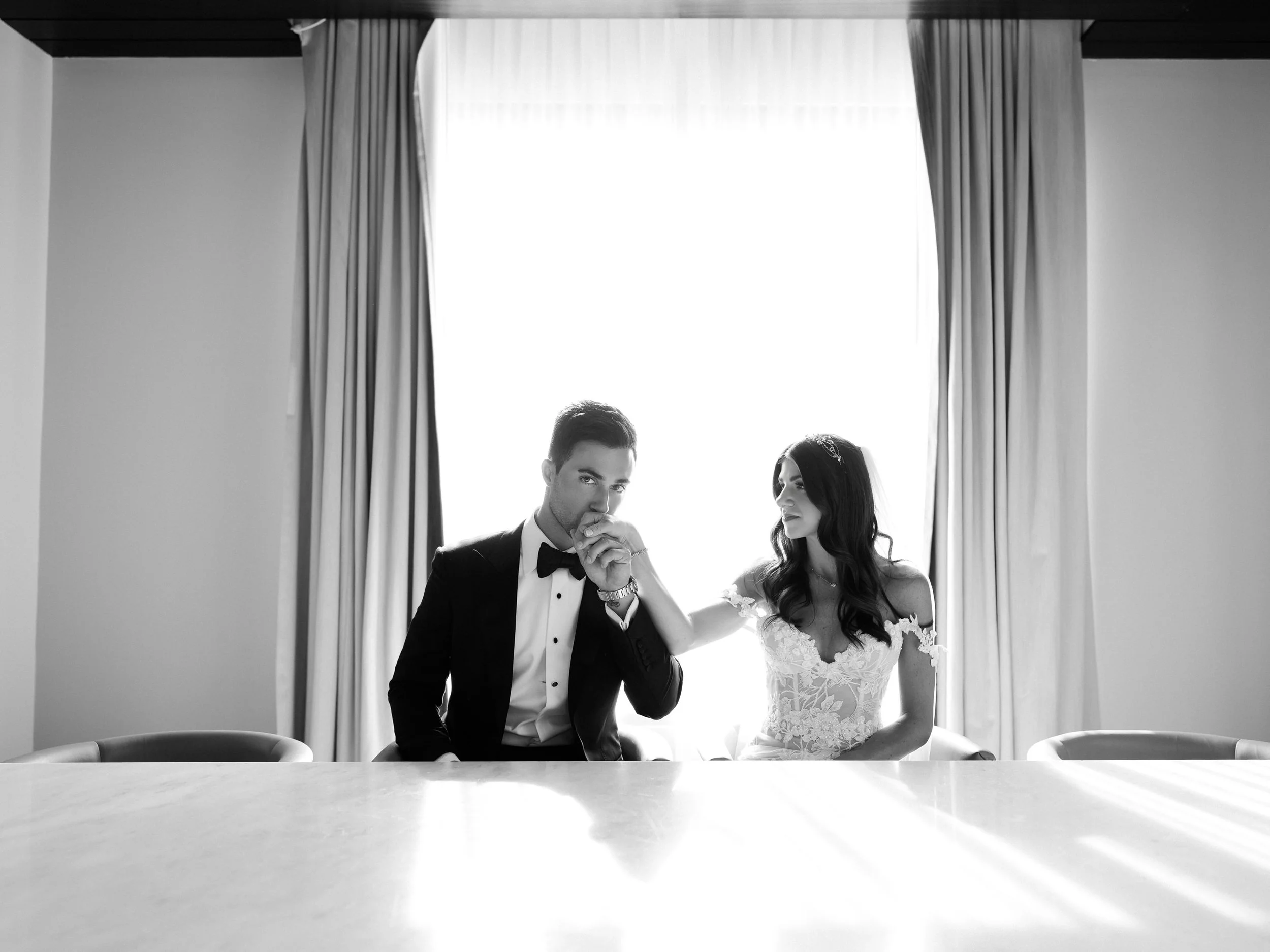 A black and white photo of a bride and groom sitting at a table. The groom, dressed in a tuxedo with a bow tie, is kissing the bride's hand. The bride is wearing a wedding dress with lace details and has long dark hair. They are in a room with a larg