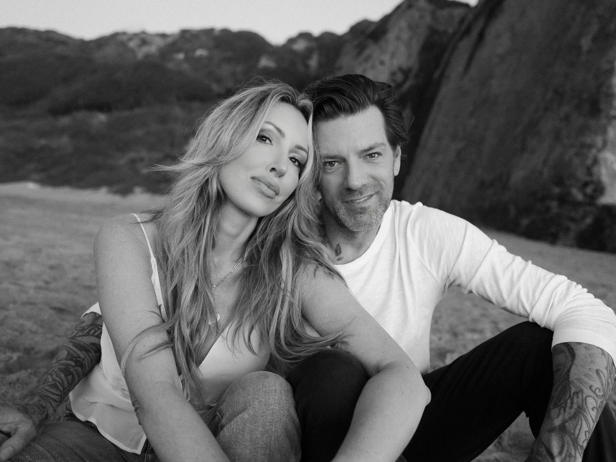 A black-and-white photo of a couple sitting outdoors on a beach with mountains in the background. The woman has long wavy hair and is wearing a sleeveless top, while the man has short hair and is wearing a white long-sleeve shirt. They are sitting cl
