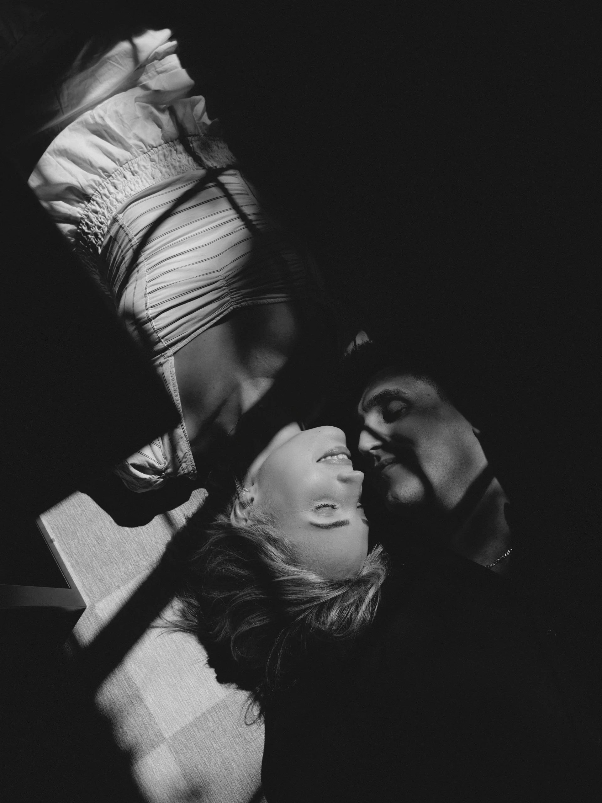A black and white photo of a young couple lying down face-to-face with eyes closed, smiling, in intimate closeness, with shadows playfully cast on their faces.