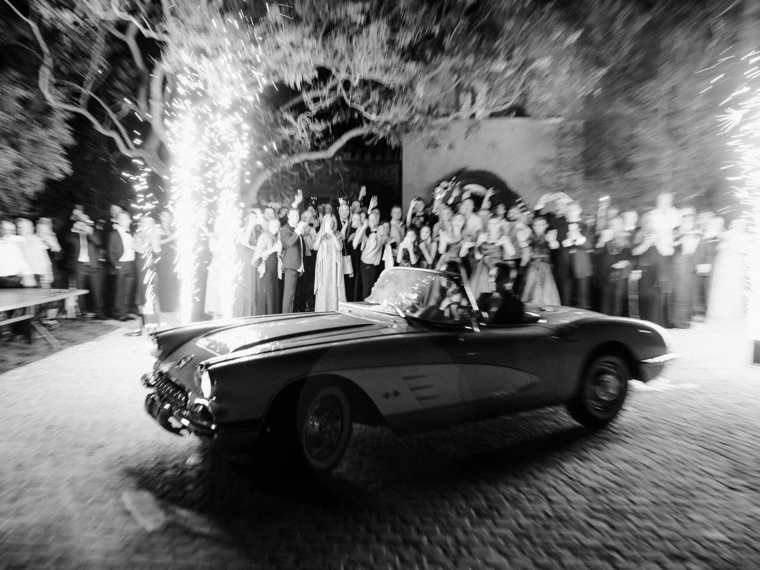 A vintage convertible car driving through a celebration, with a large crowd of people and sparklers lighting up the scene at night.