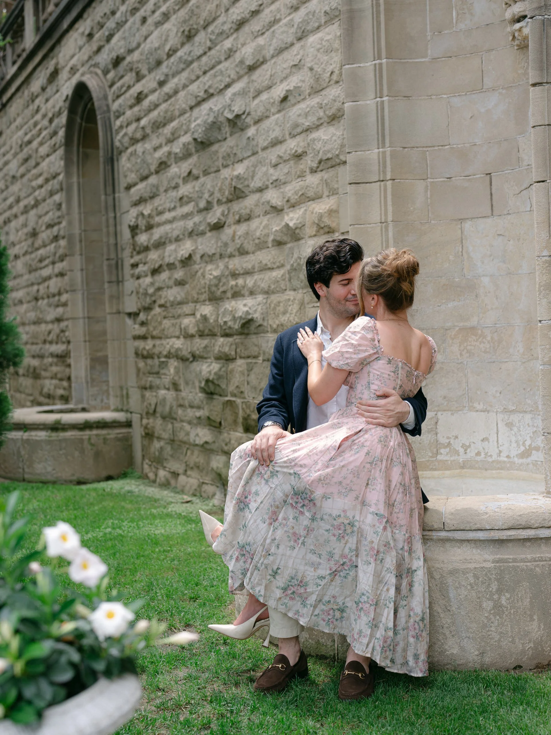 A man and woman dance outdoors near a stone building, with the woman lifting her leg in a floral dress and high heels, and the man in a navy blazer and brown shoes, embracing and smiling at each other.