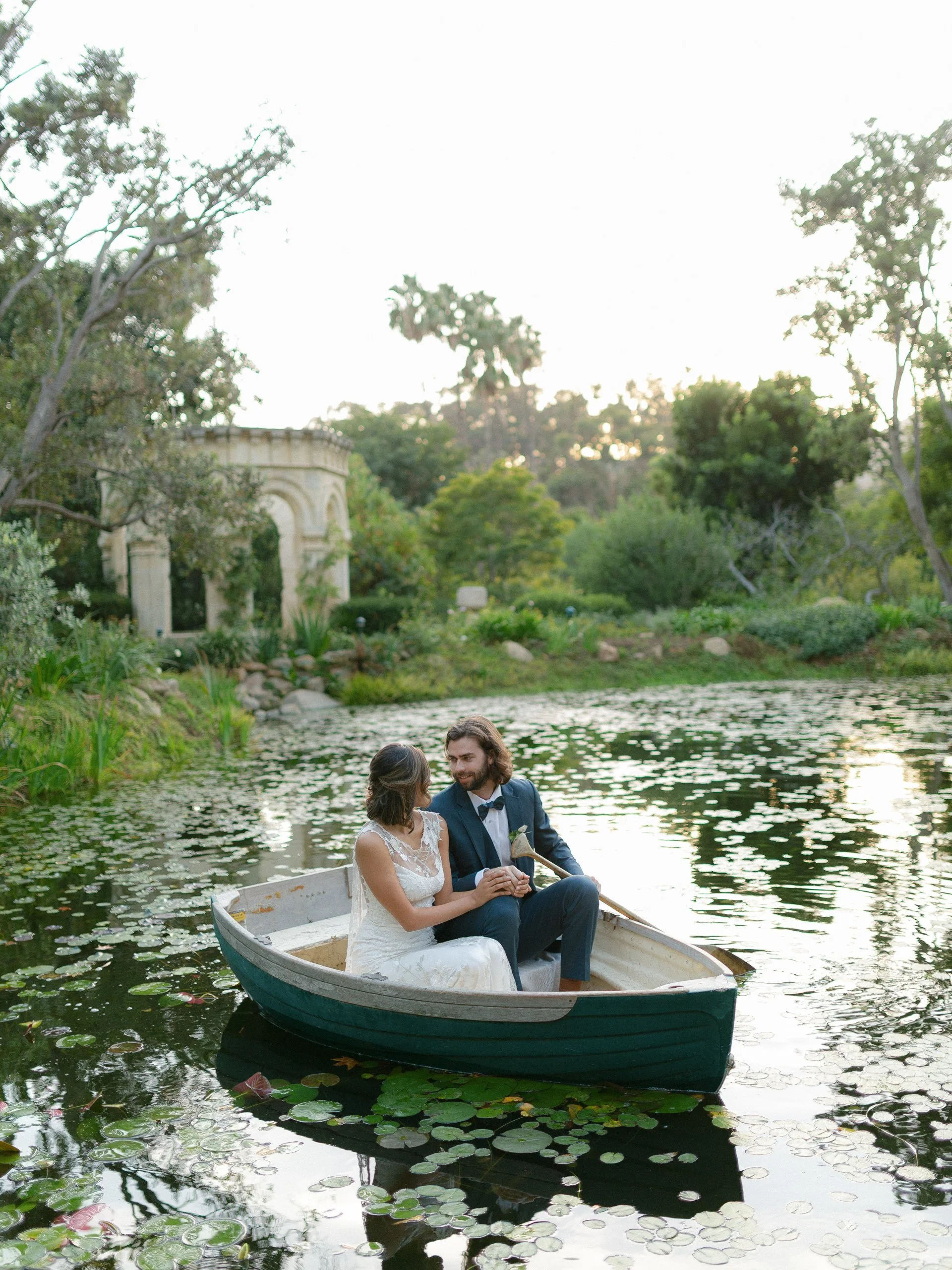 A couple in wedding attire sitting in a rowboat on a pond surrounded by lily pads, with trees and a small architectural structure in the background during sunset.
