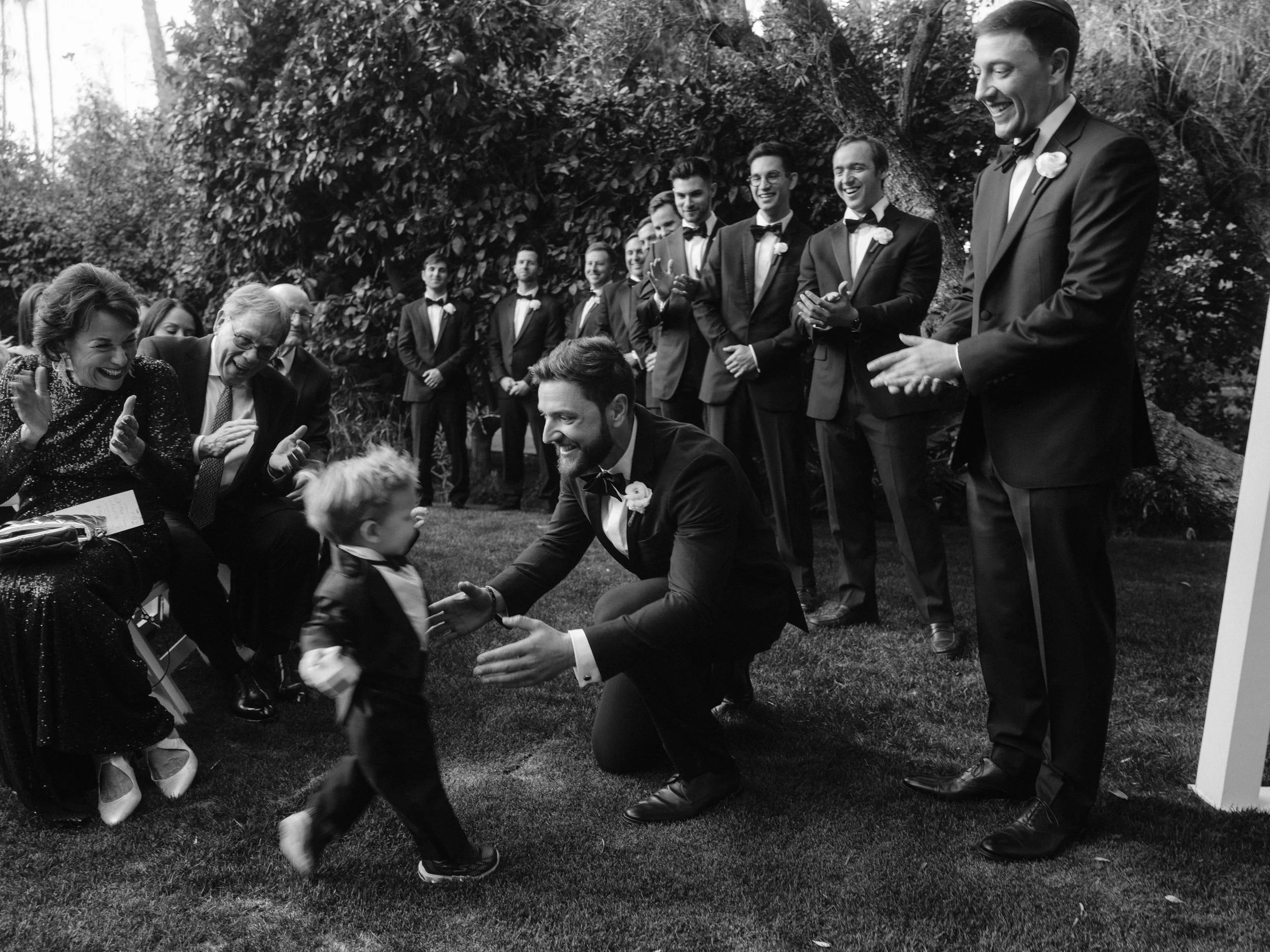 A black and white photo of a wedding celebration outdoors. A man in a tuxedo kneels on the grass, happily interacting with a young boy, while a group of men in suits and women in elegant attire watch and cheer.