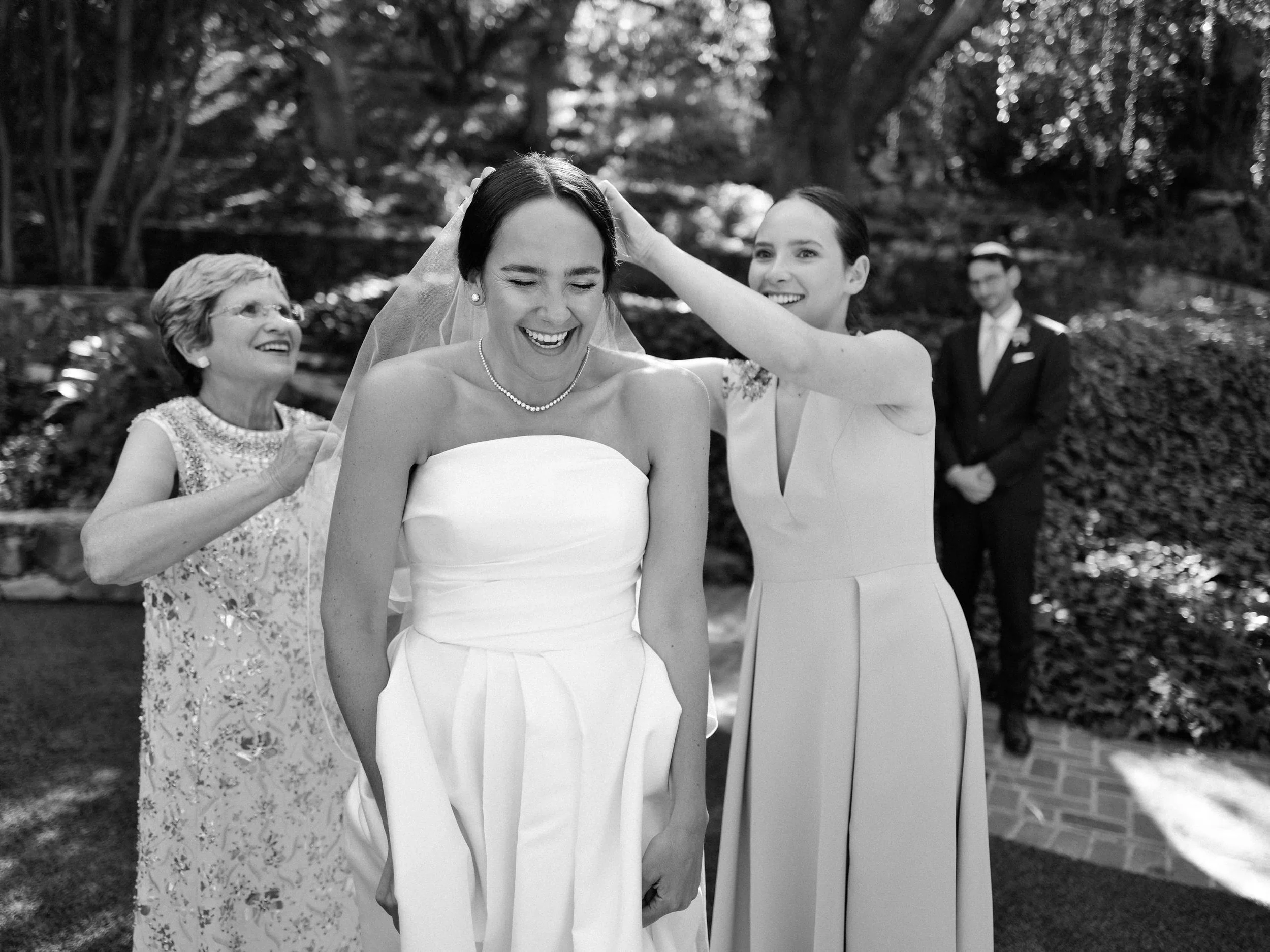 Black and white photo of a wedding scene with four women, including a bride in a strapless wedding dress being helped with her veil by a woman in a sleeveless dress, an older woman in a floral dress smiling behind them, and a man in a suit and tie in