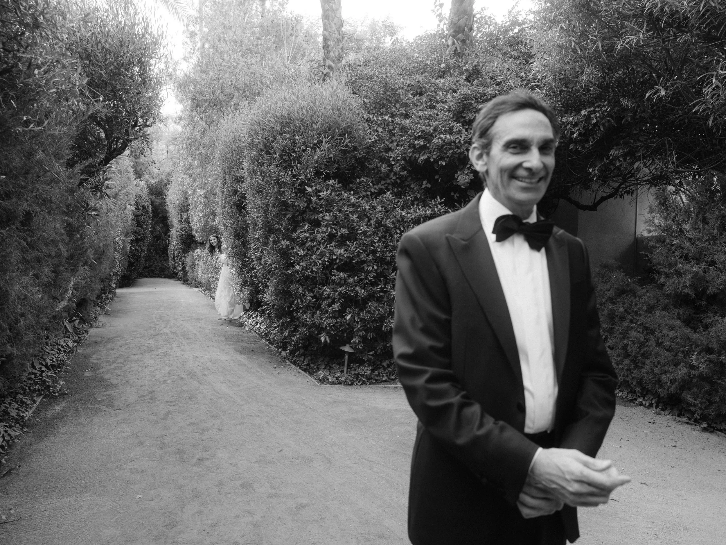 A man in a tuxedo with a bow tie smiling in the foreground, a woman in a wedding dress standing on a pathway surrounded by trees and foliage in the background.