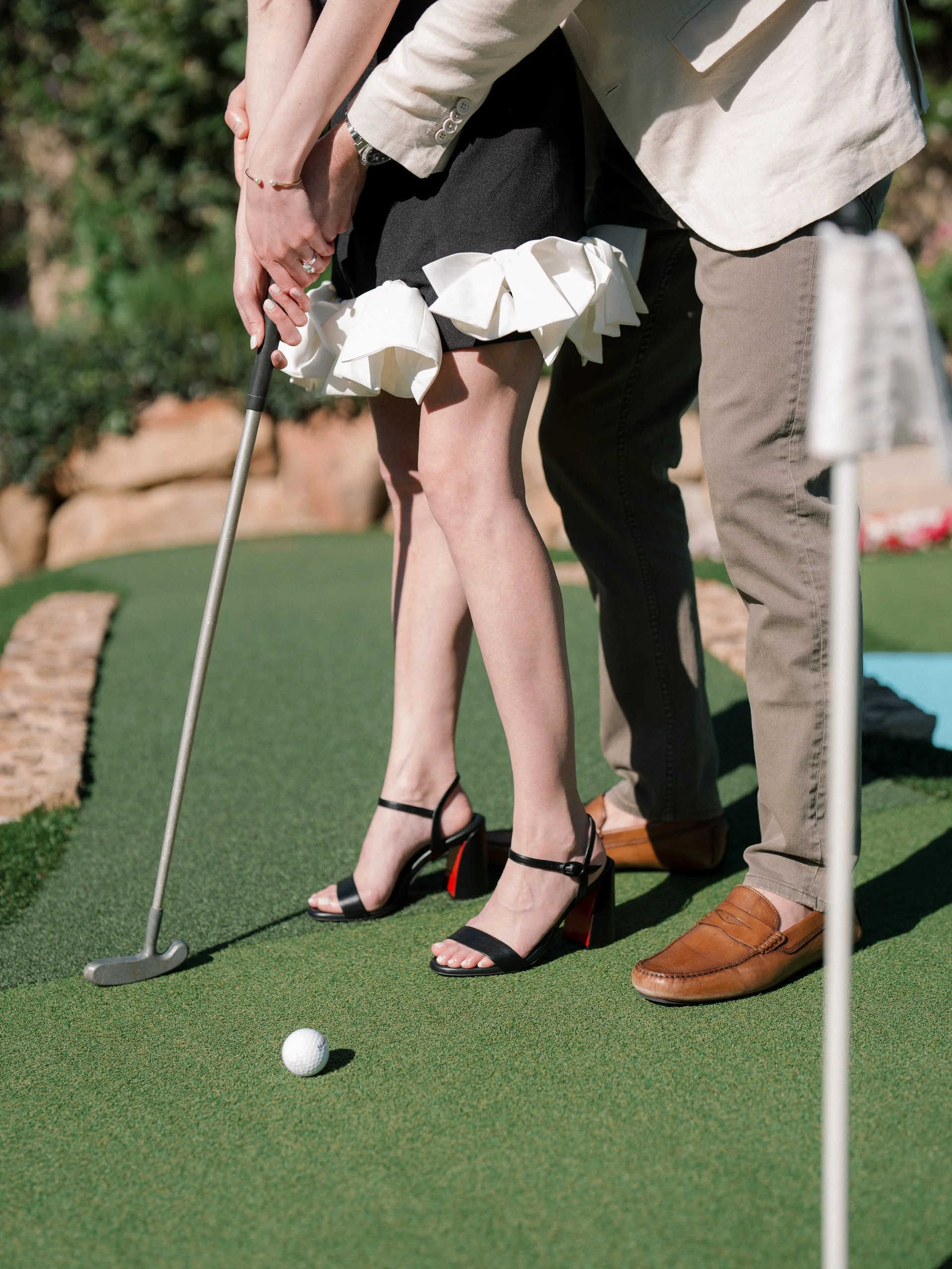 A woman in black high heels is putting a golf ball on the putting green with help from a man in brown shoes during a mini golf game.