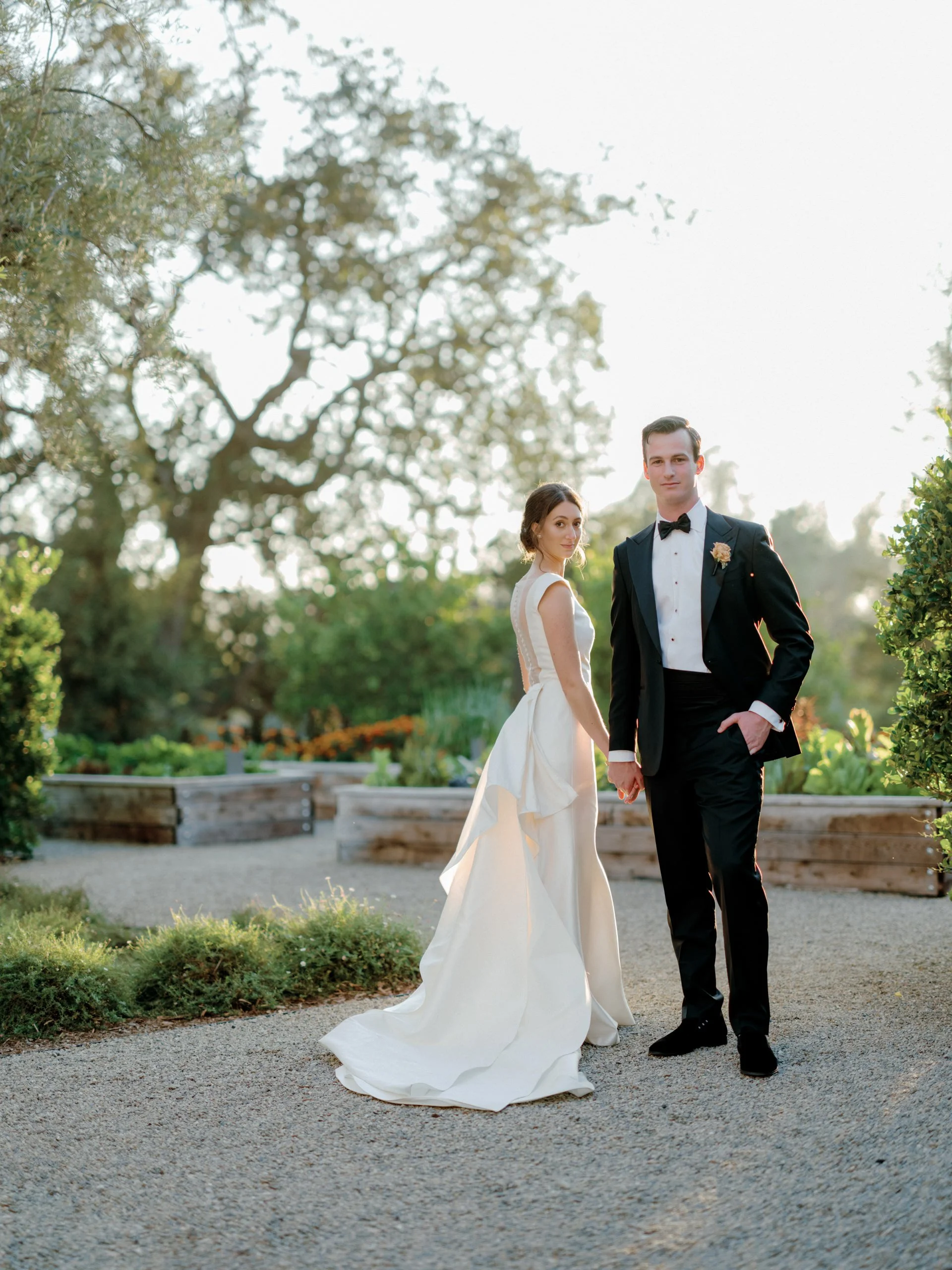 A bride and groom holding hands outdoors during sunset, with trees and plants in the background.