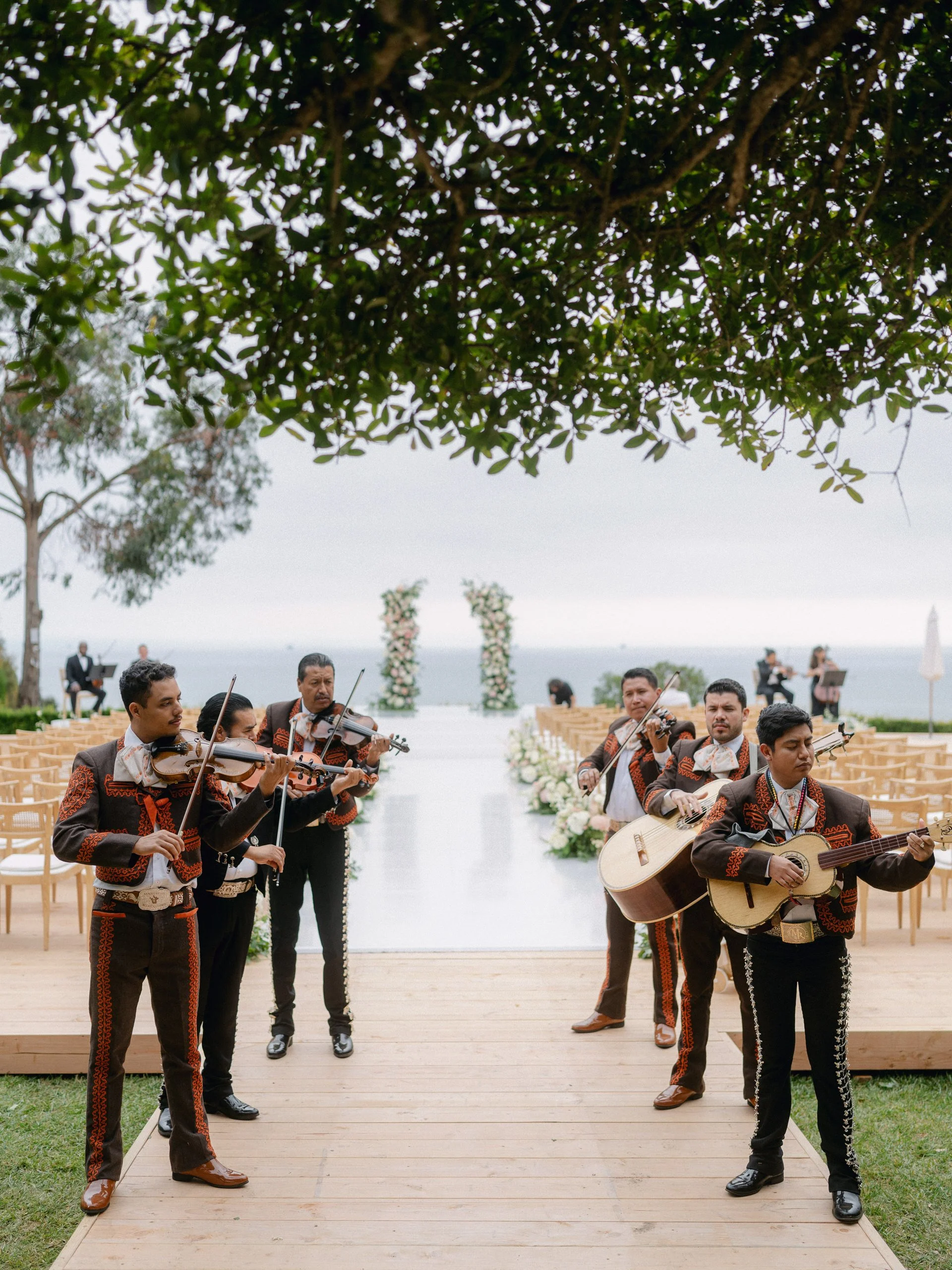 Mariachi band playing music at outdoor wedding ceremony on a wooden aisle with floral arches and ocean in the background.