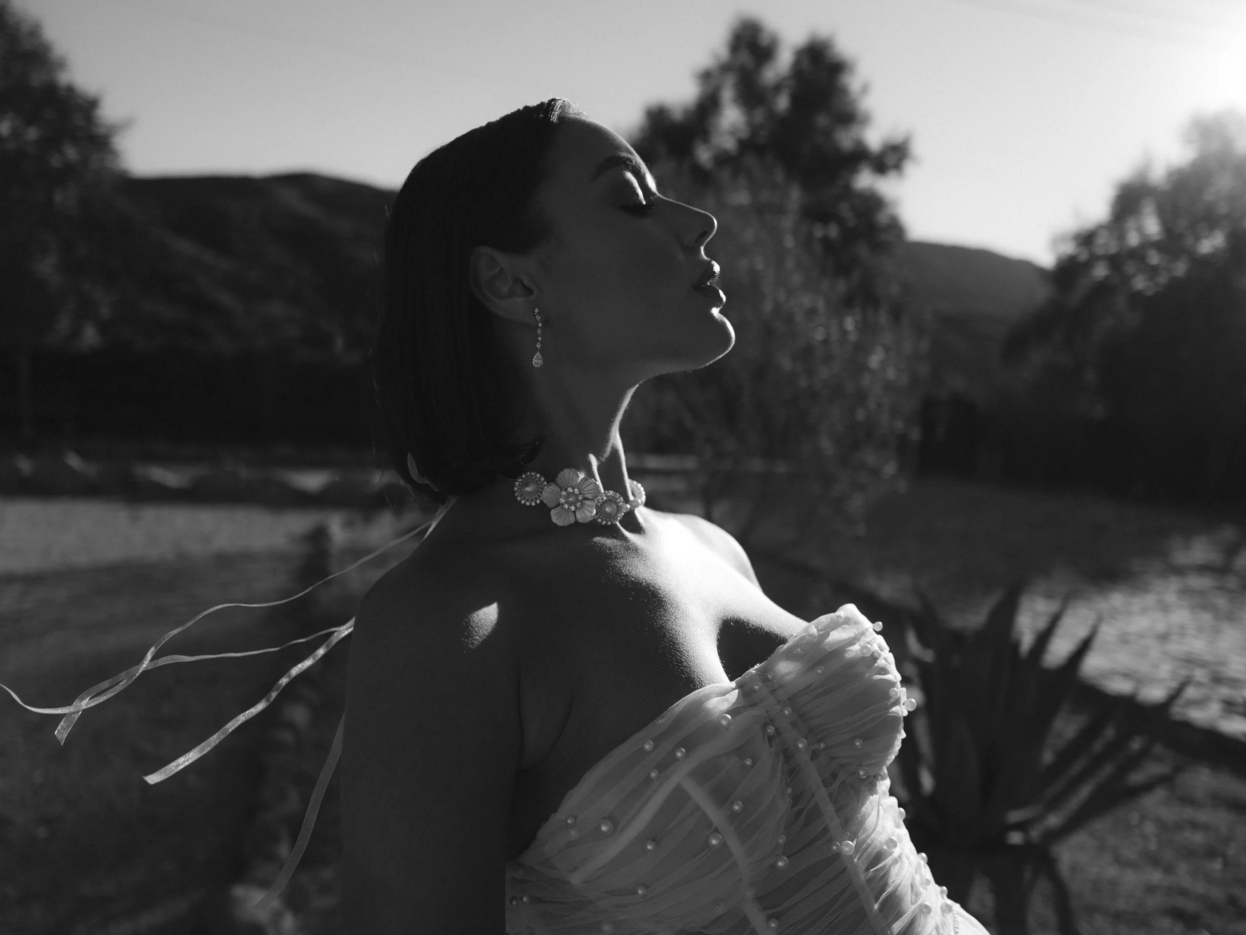 A woman with short hair and jewelry, wearing an off-shoulder dress, stands outdoors with her eyes closed, facing the sun, with trees and hills in the background.