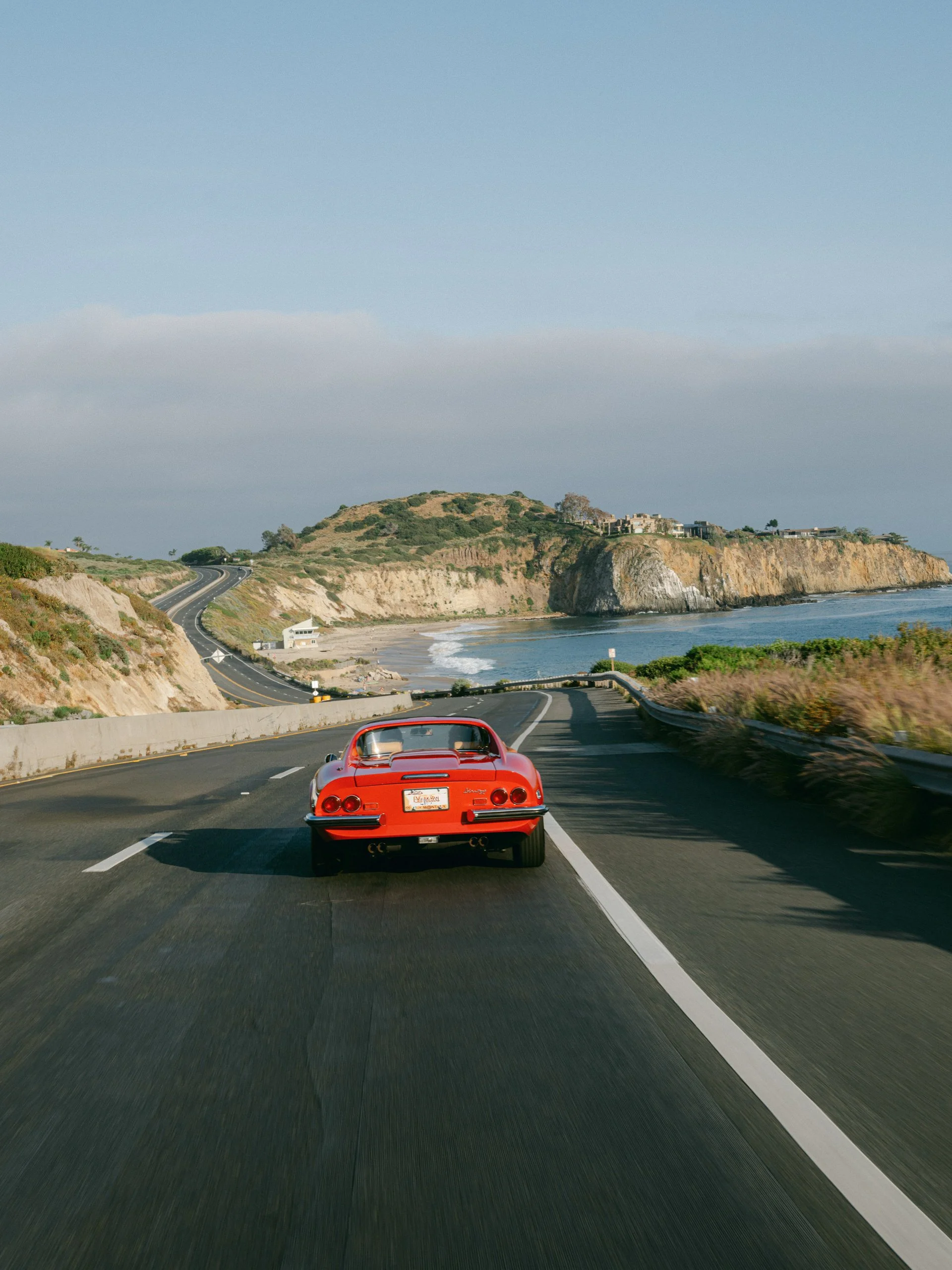 Red vintage sports car driving on a coastal highway with cliffs and ocean views.