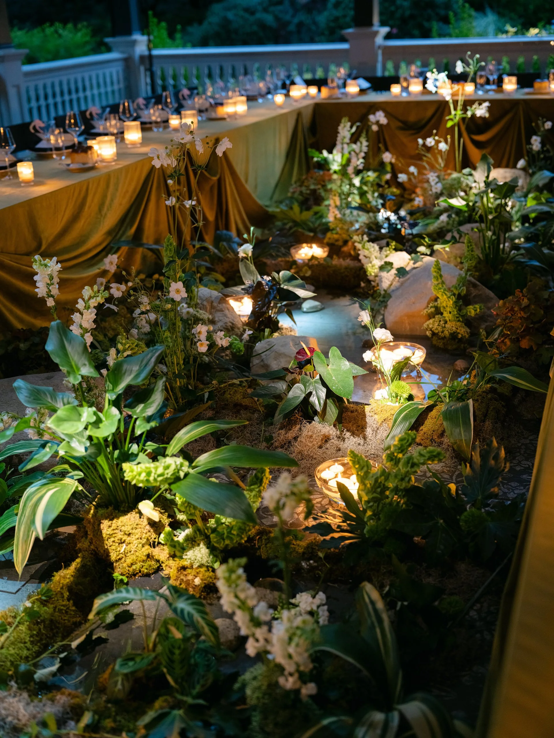 A long dining table decorated with candles, flowers, and draped fabric, set outdoors on a porch or balcony with a view of trees.