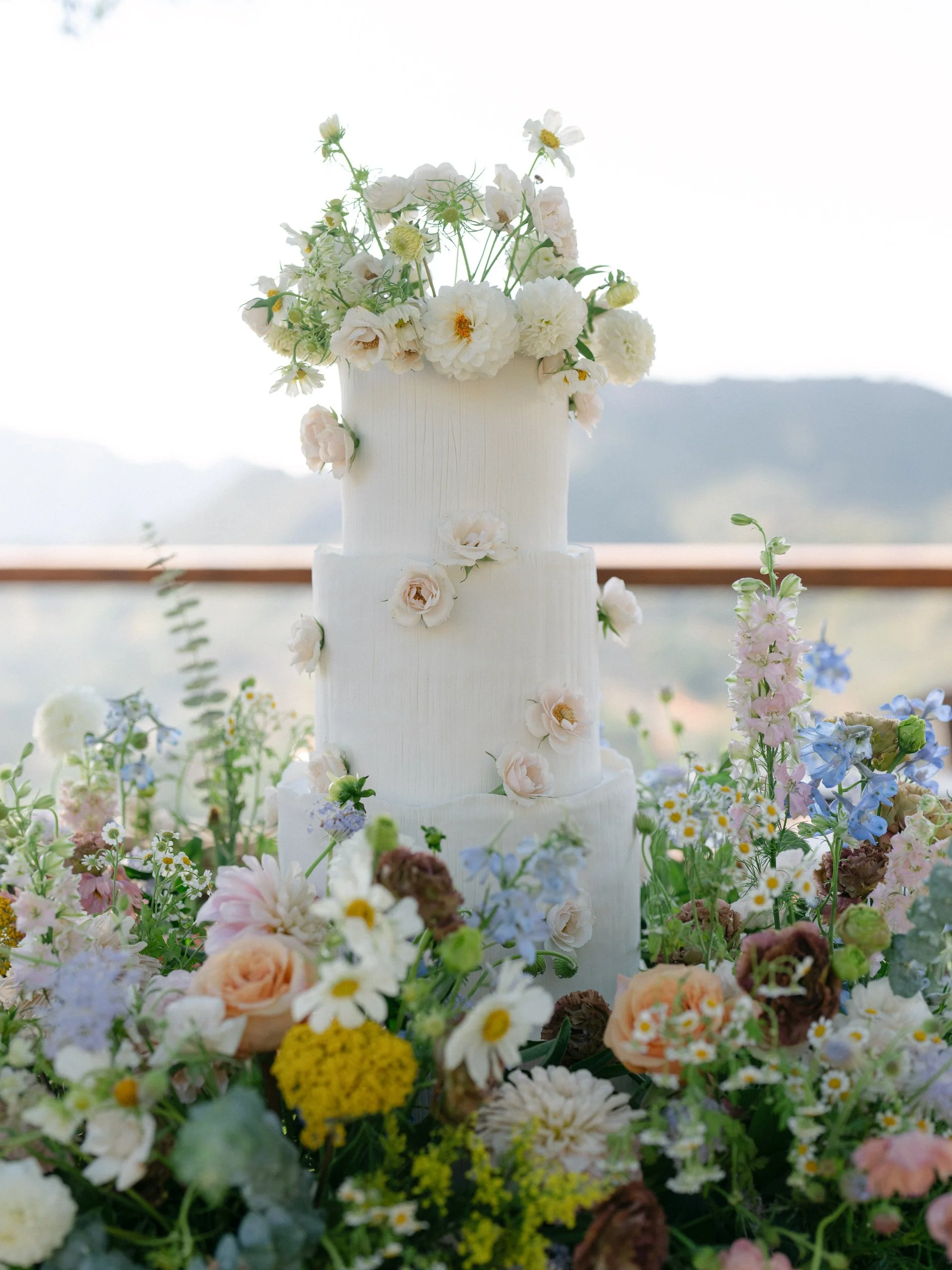 A tiered white wedding cake decorated with white and blush flowers, surrounded by a colorful assortment of mixed flowers.