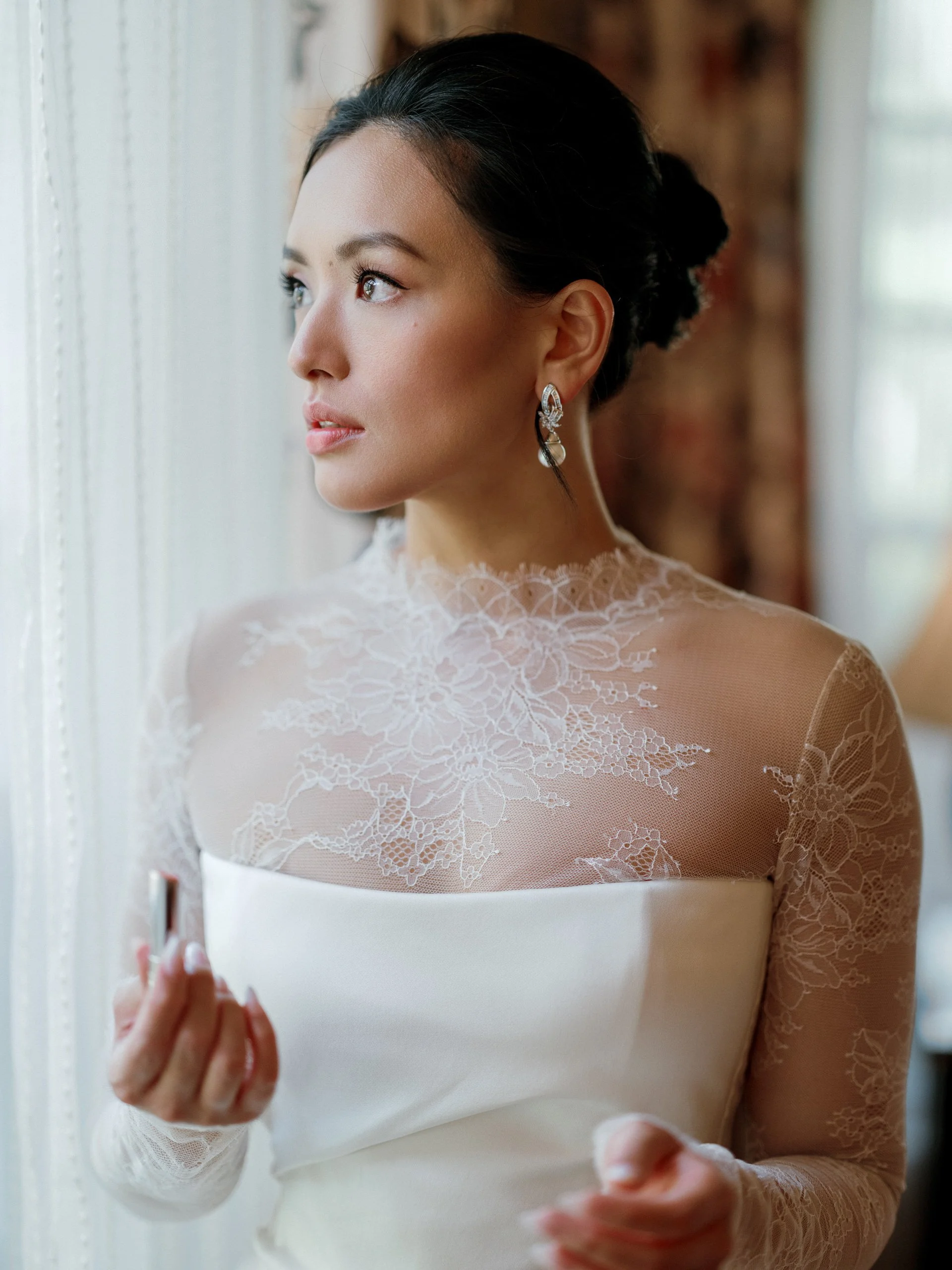 A woman in a white wedding dress with lace details, wearing earrings, looking out a window